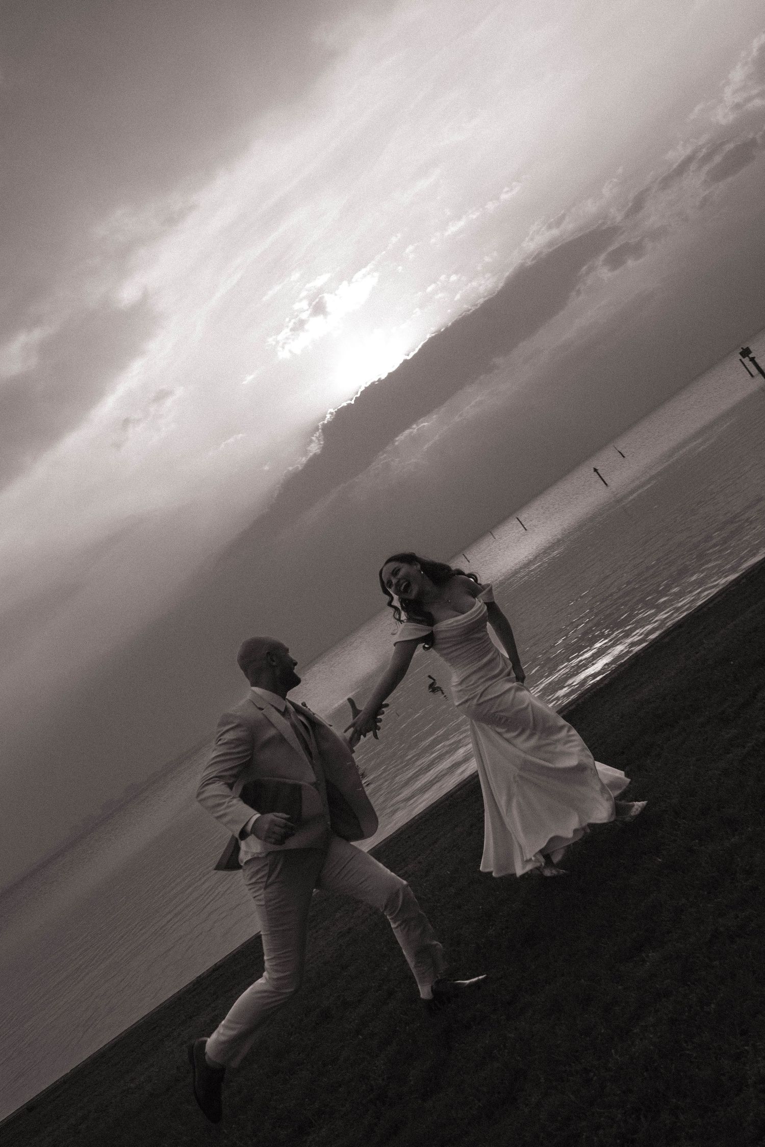 Black and white photo of joyful couple running on beach at dramatic sunset by ocean.