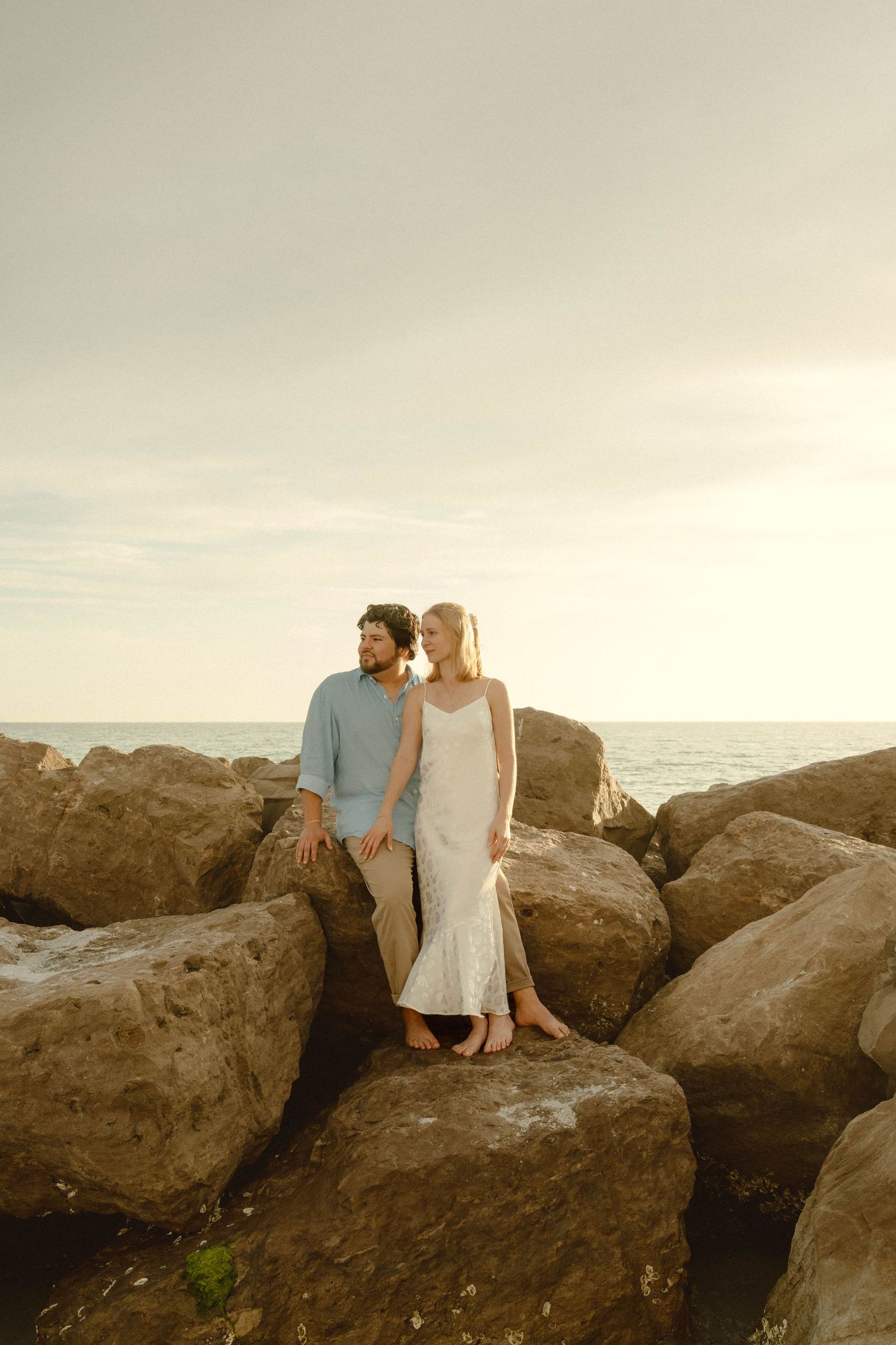 Couple standing together on large rocks by the ocean during golden hour sunset.