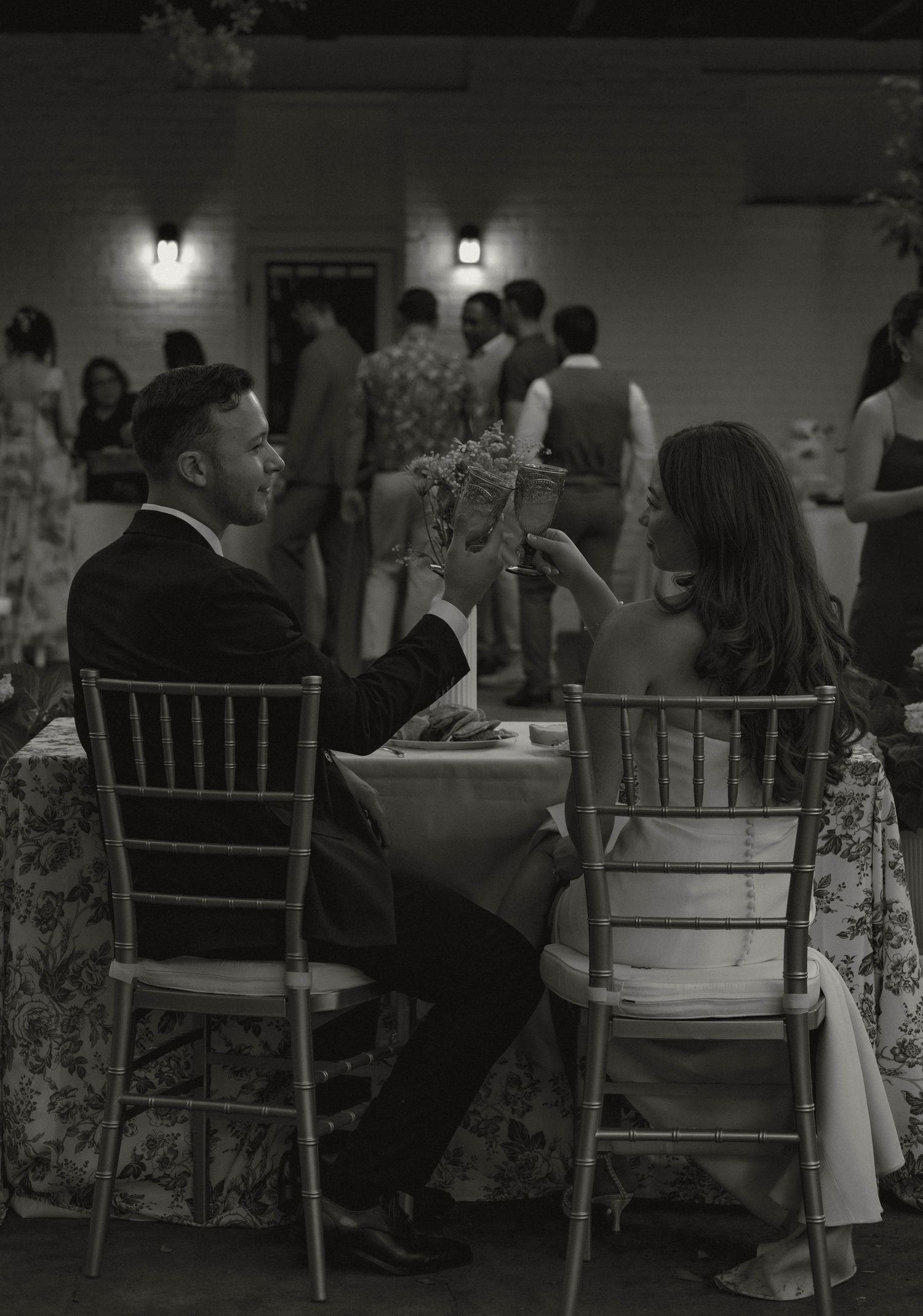 Black and white photo of bride and groom toasting at wedding reception table.