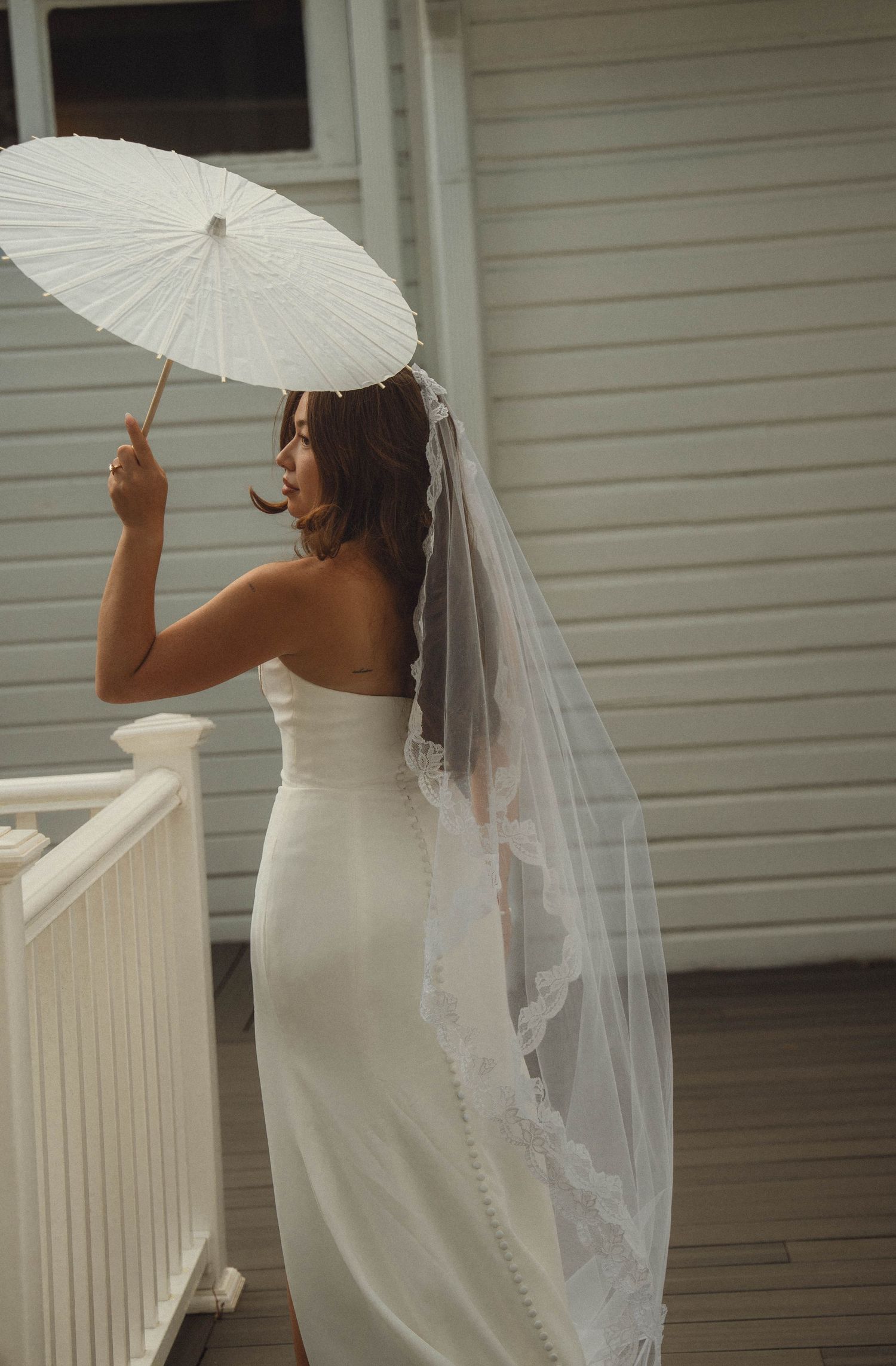 Bride in strapless white gown and cathedral veil holds a white parasol on a wooden porch.