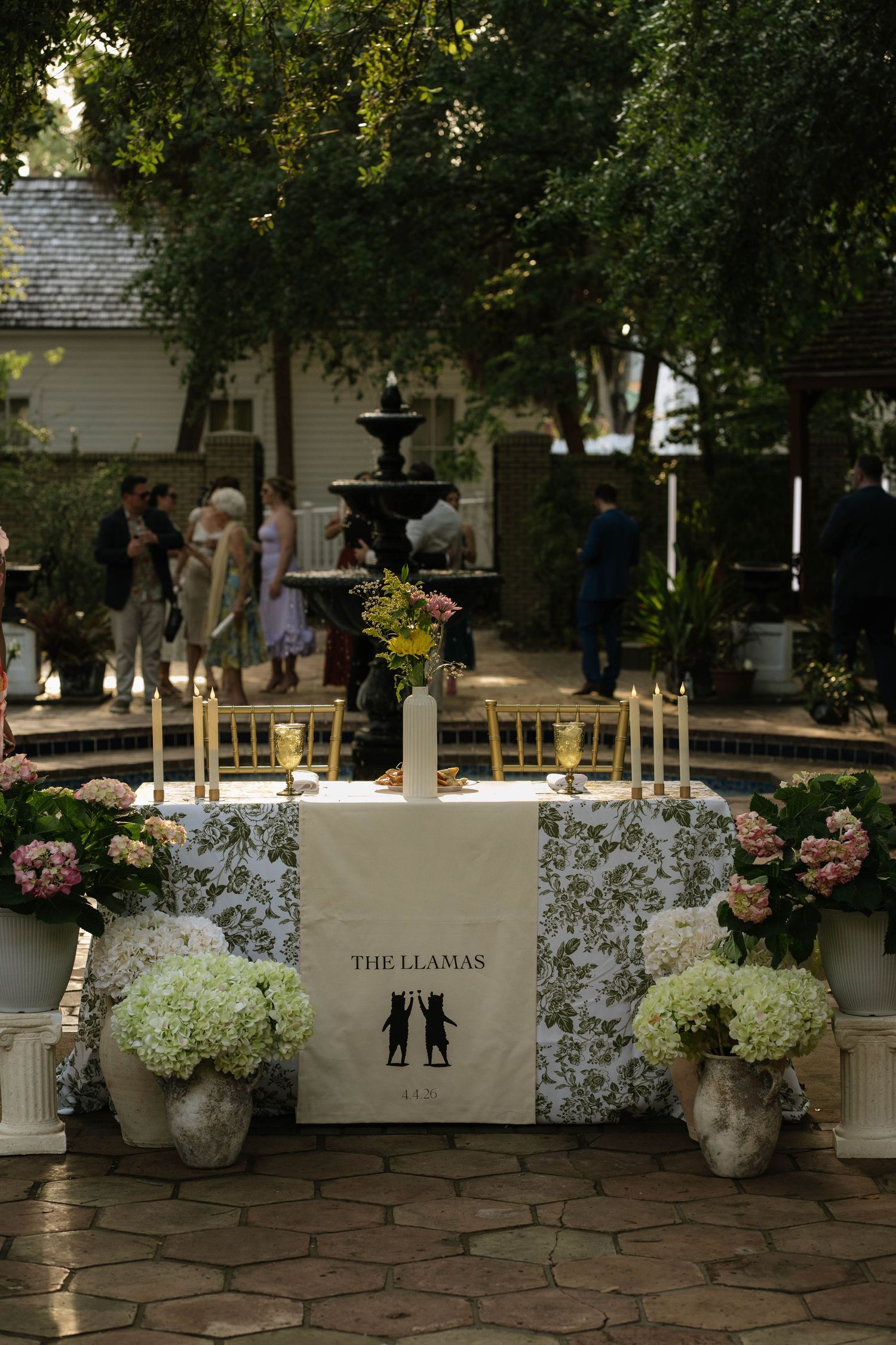Outdoor wedding sweetheart table decorated with hydrangeas and candles near a garden fountain.