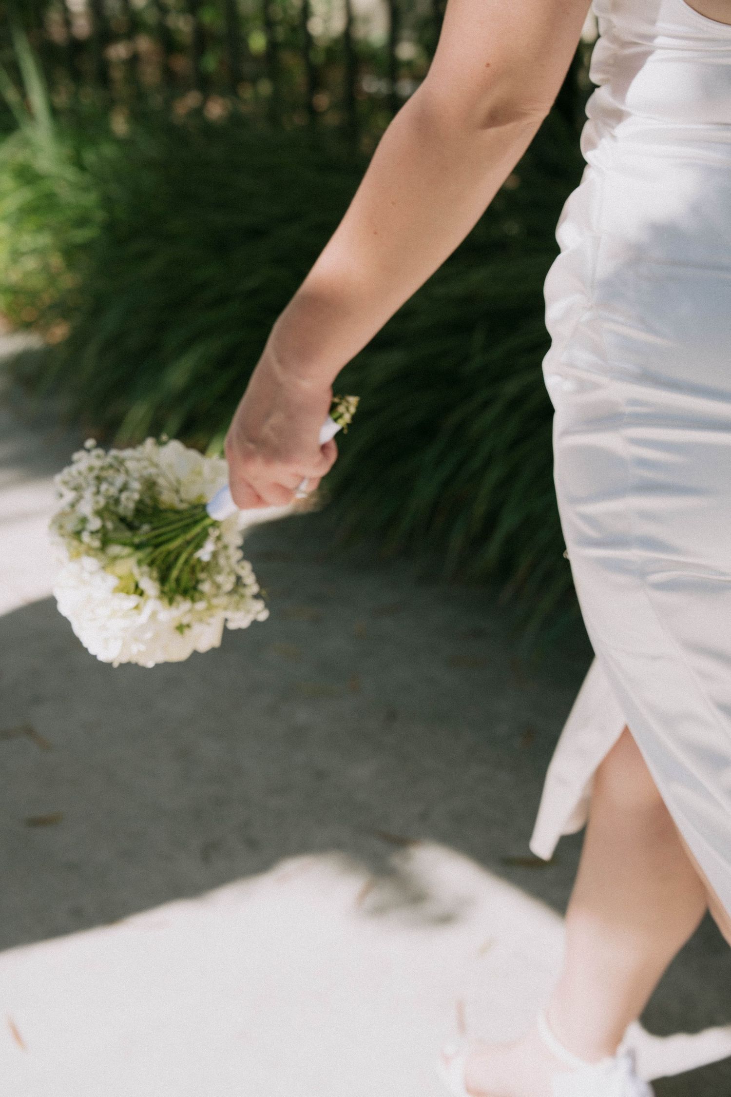 Close-up of bride in white dress carrying small white floral bouquet while walking outdoors.