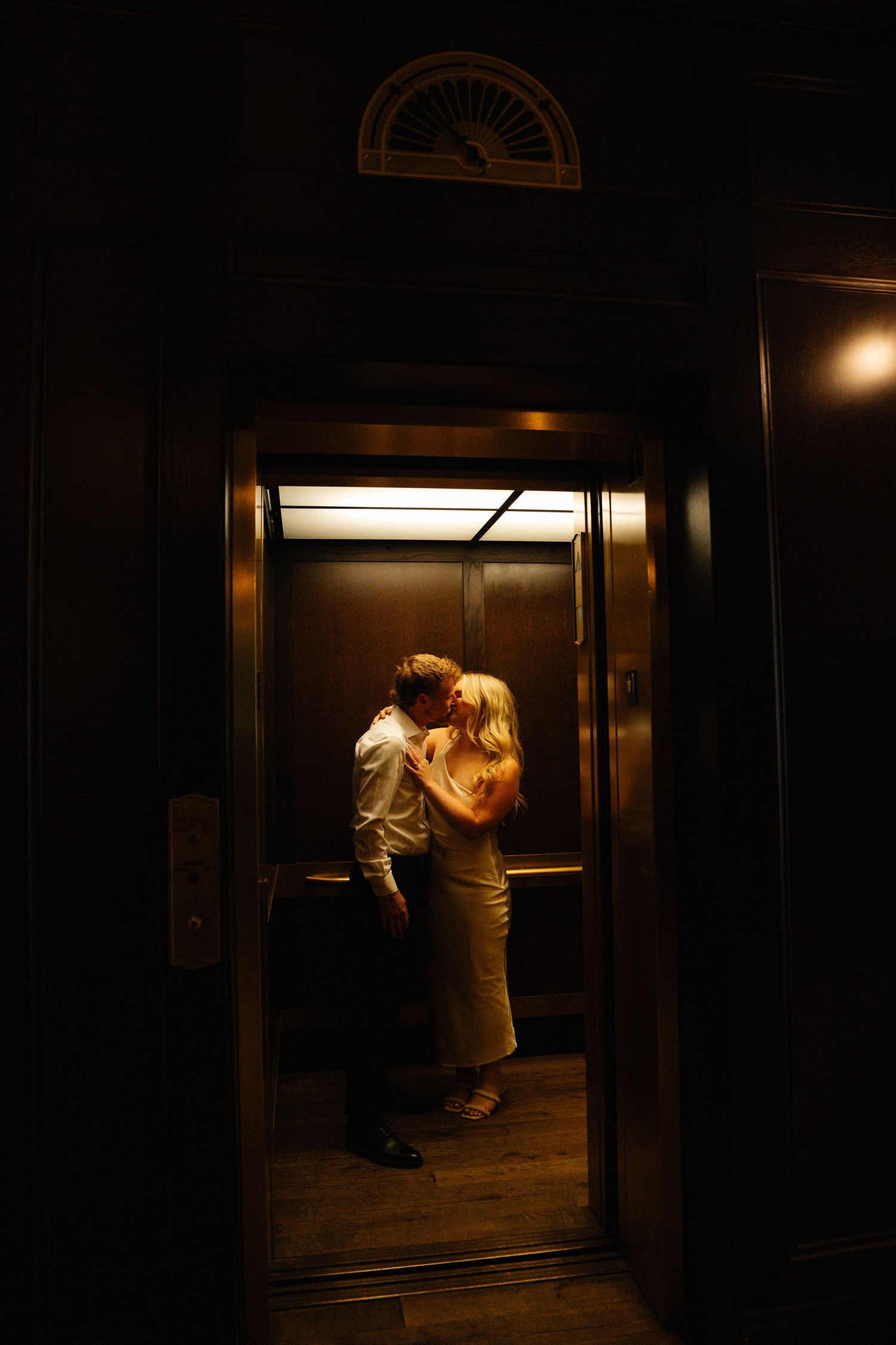 Couple sharing a romantic kiss inside a warmly lit elevator viewed through open doors at night.