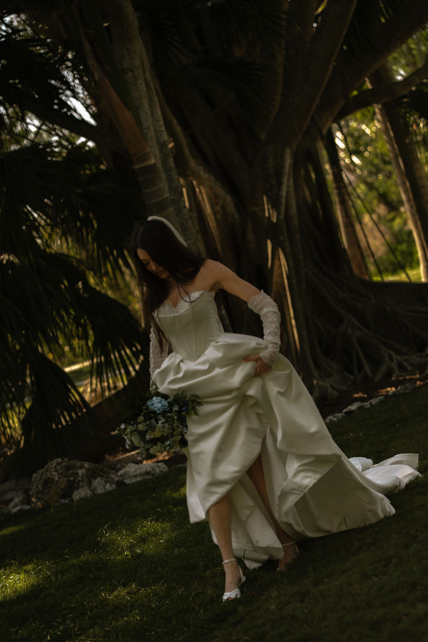 Bride in white strapless gown holding blue bouquet walks gracefully beneath a large banyan tree in lush garden.