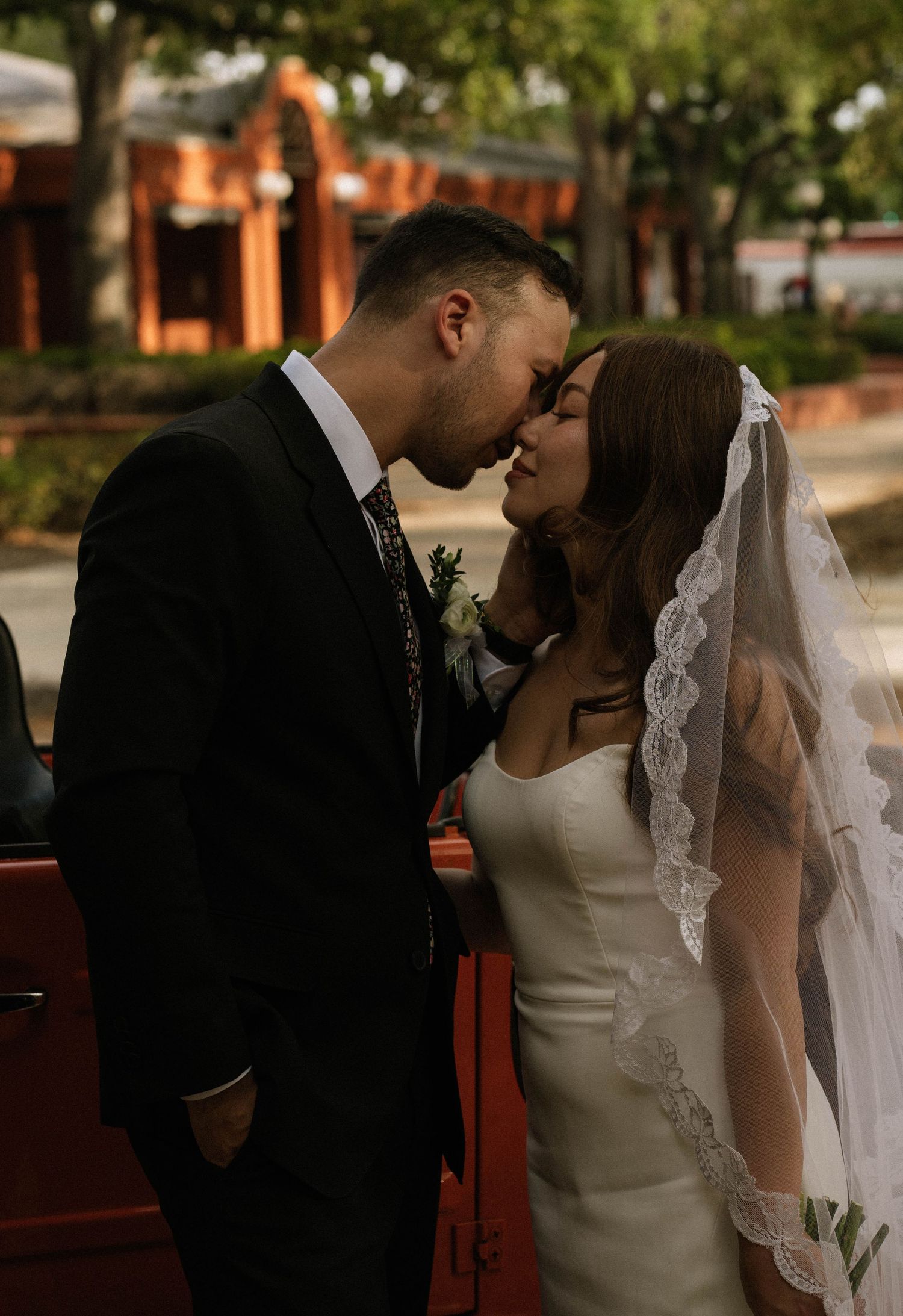 Newlyweds share a tender almost-kiss beside a red vintage car, bride wearing lace-edged veil.