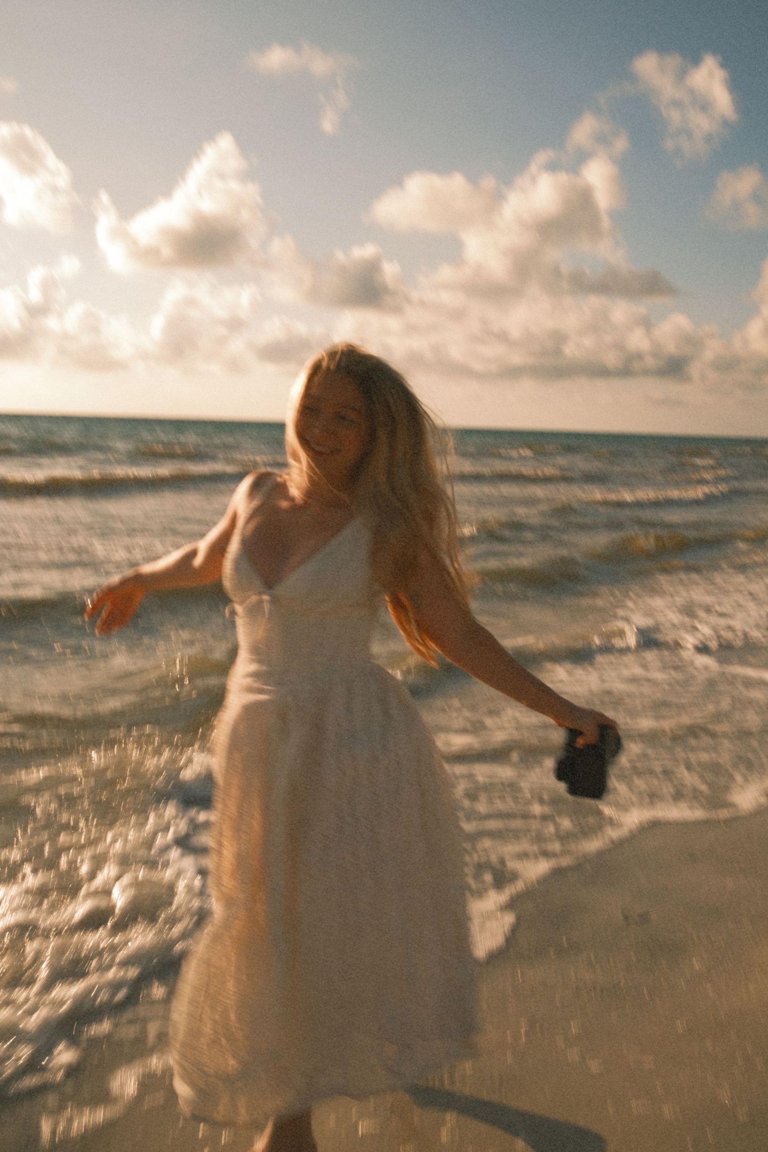 A woman in a white dress walks along a sunny beach, holding a camera as waves wash over her feet.