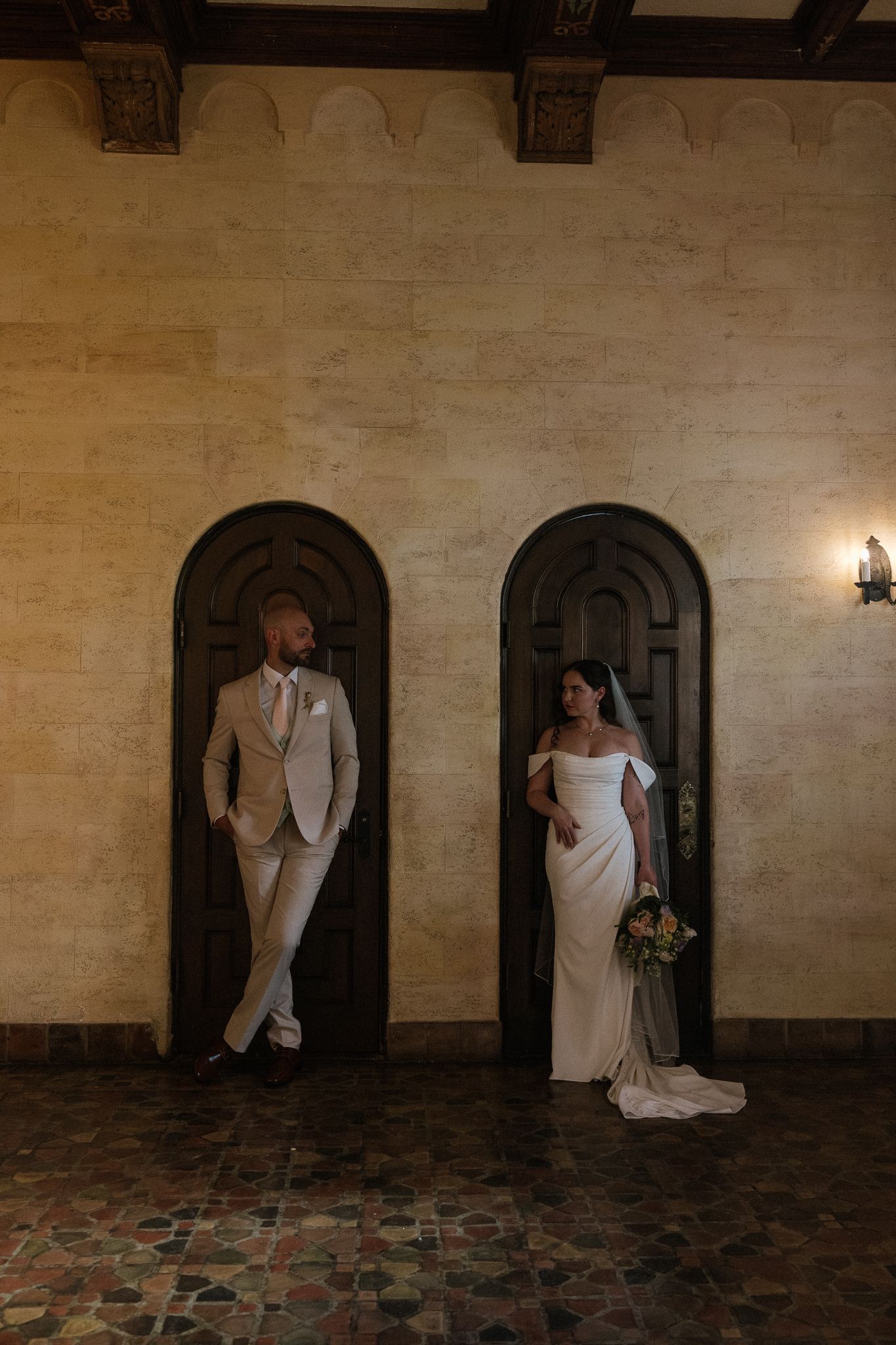 Bride and groom pose in separate arched doorways in a warmly lit stone hall, bride holding a bouquet.