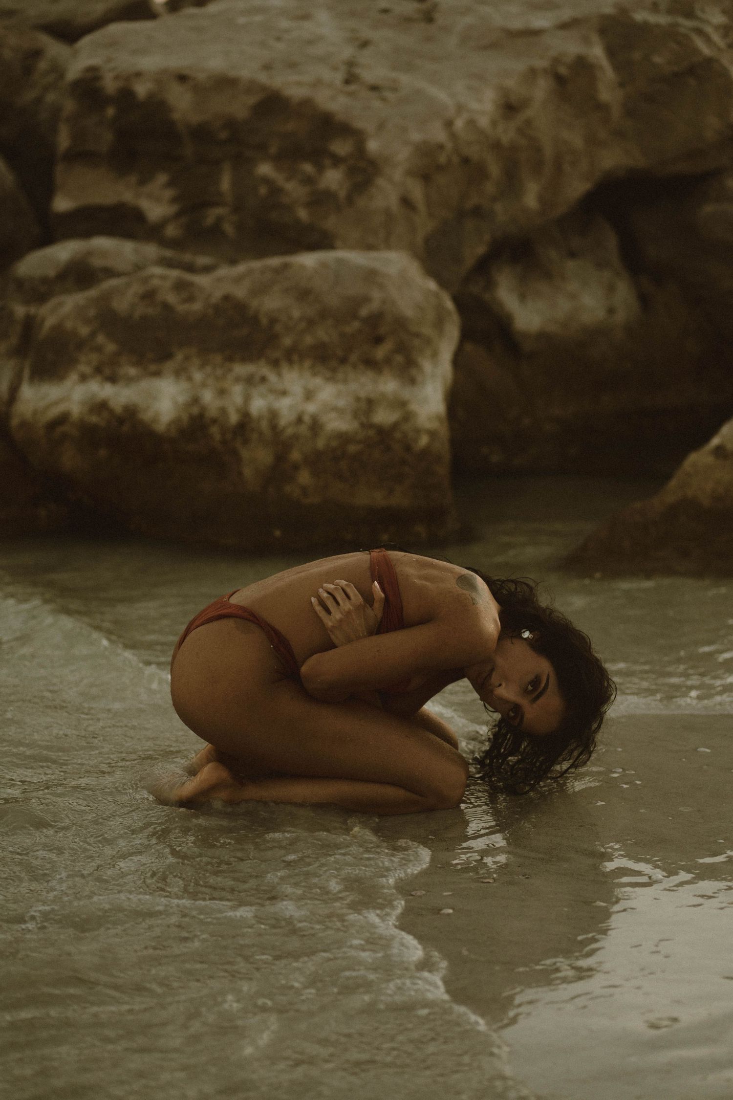 Woman in red bikini crouching in shallow ocean water with rocky backdrop.