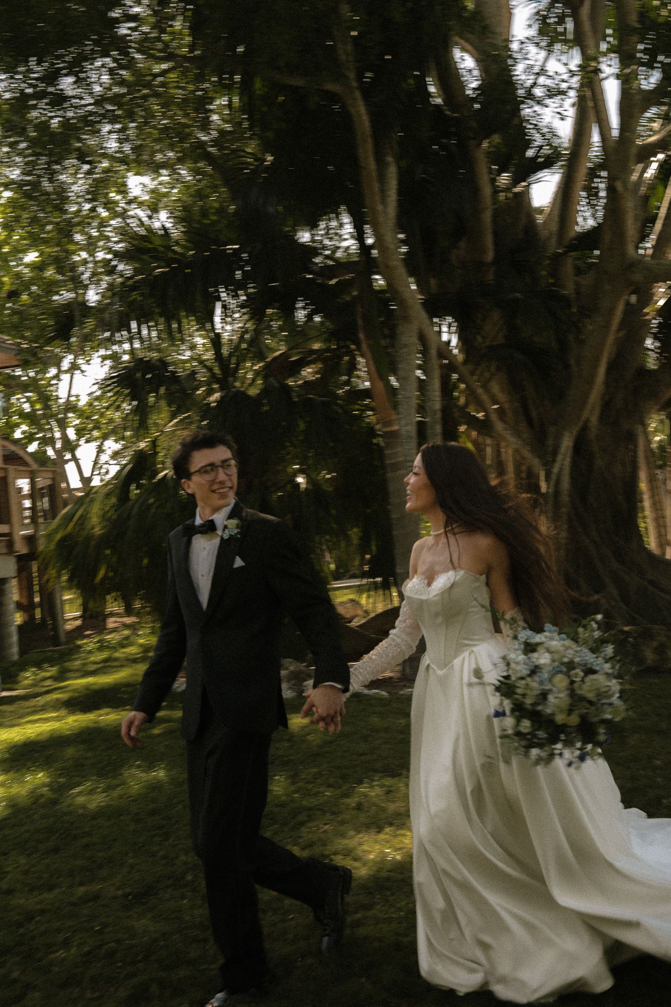 A bride and groom walk hand-in-hand through a lush green garden, she in a white gown holding a bouquet, he in a black tuxedo.