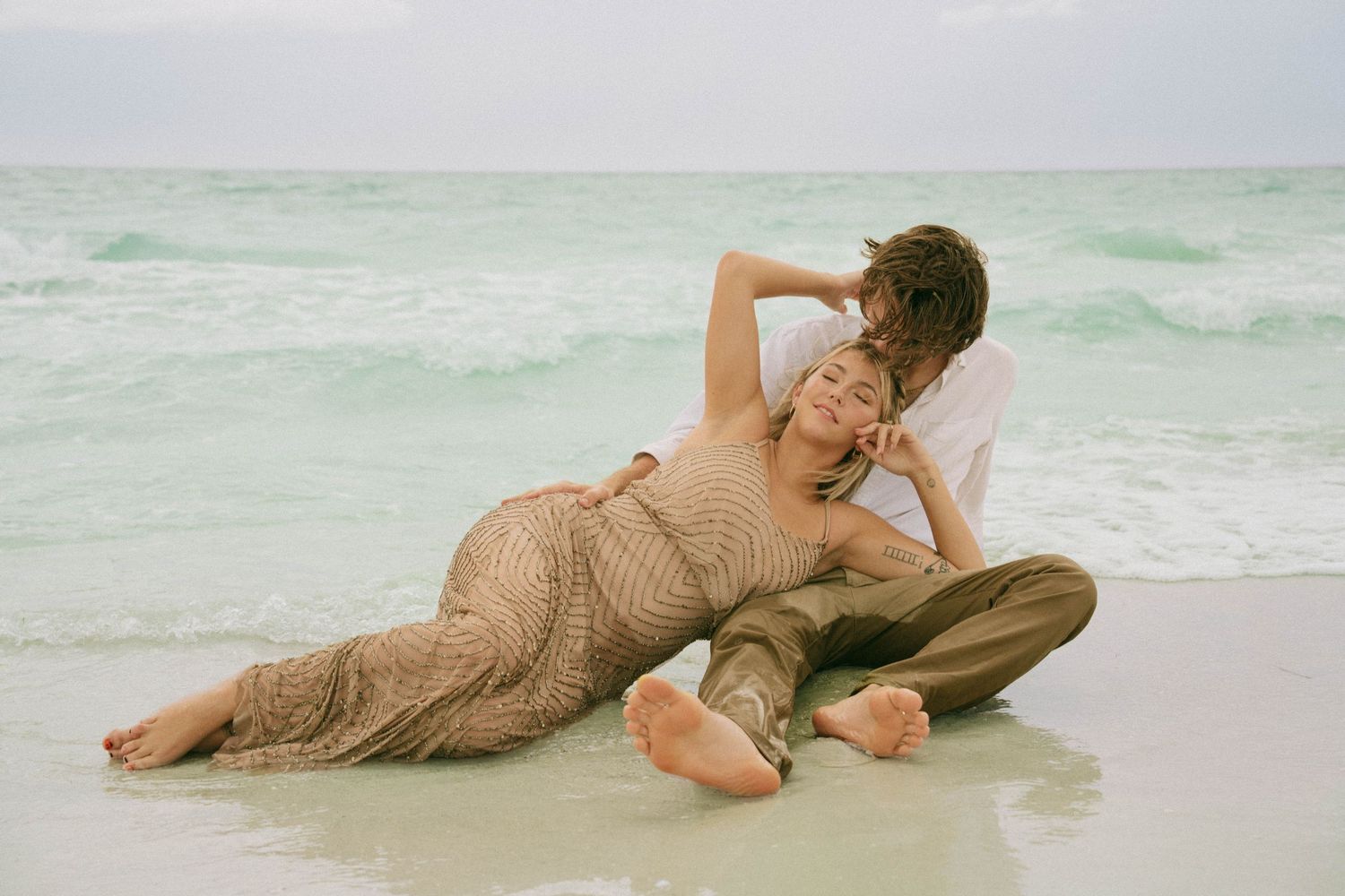 A couple poses romantically on a beach, the woman in a gold gown leaning back against her partner.