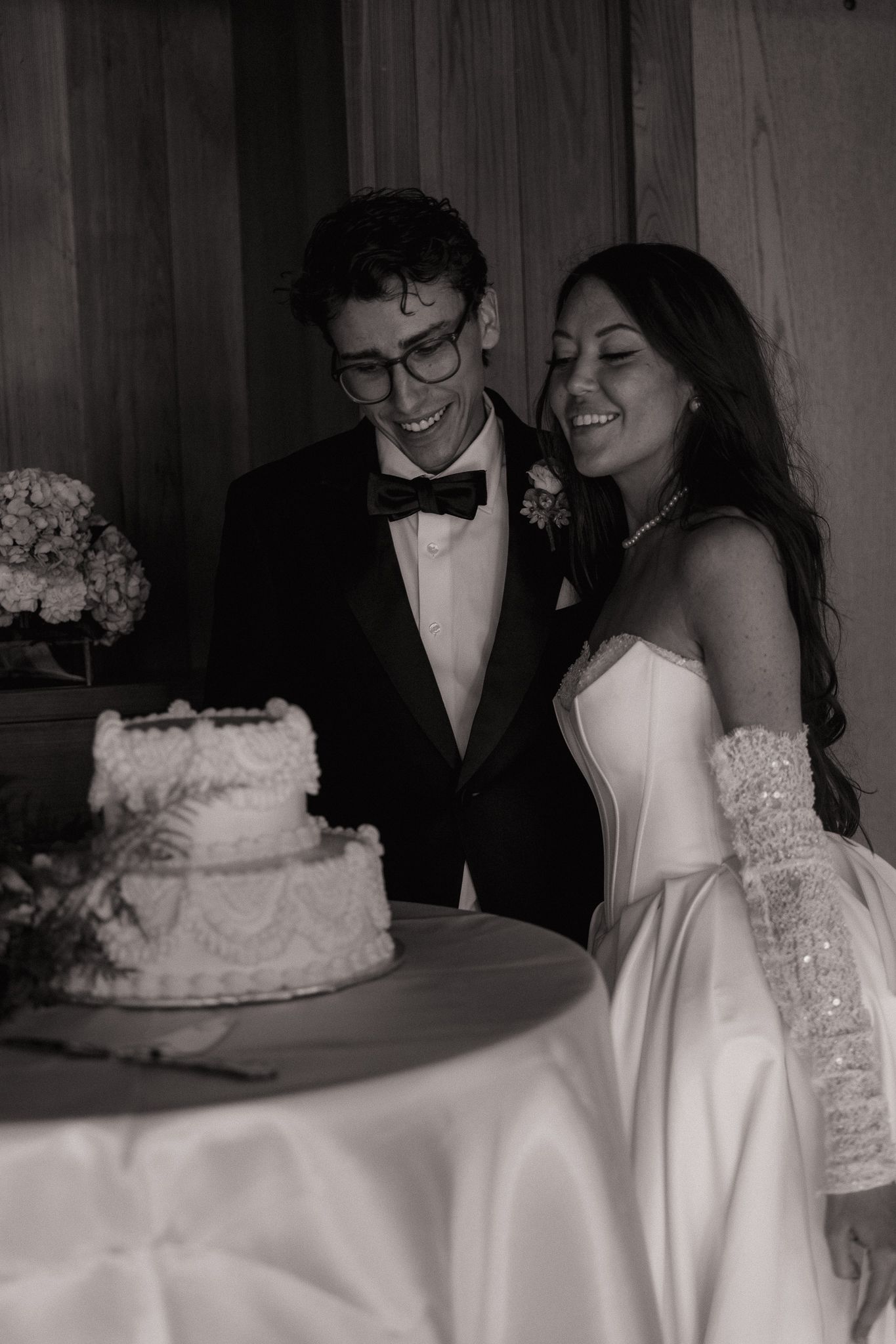 Black and white photo of a smiling bride and groom cutting their wedding cake, bride in strapless gown with gloves.
