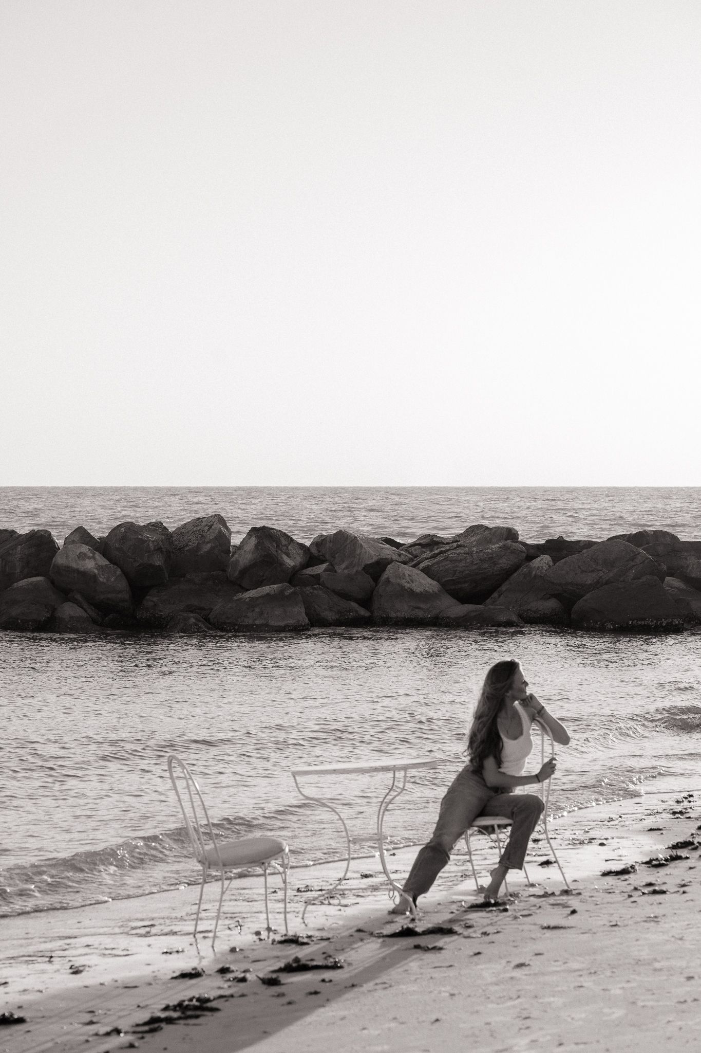 Black and white photo of woman relaxing by white metal table on sandy beach with rock jetty.
