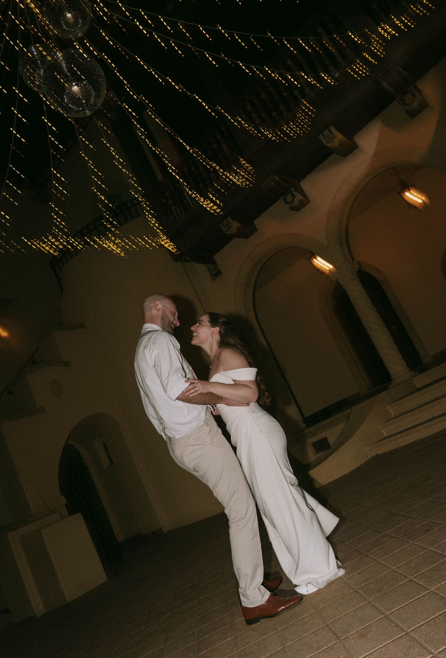 Groom lifts bride during their first dance under a canopy of twinkling string lights in an arched ballroom.