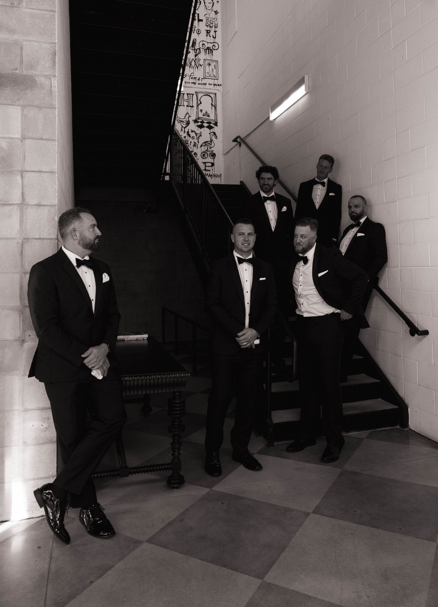 Groom and groomsmen in black tuxedos pose on a staircase in a dramatic black and white wedding photo.