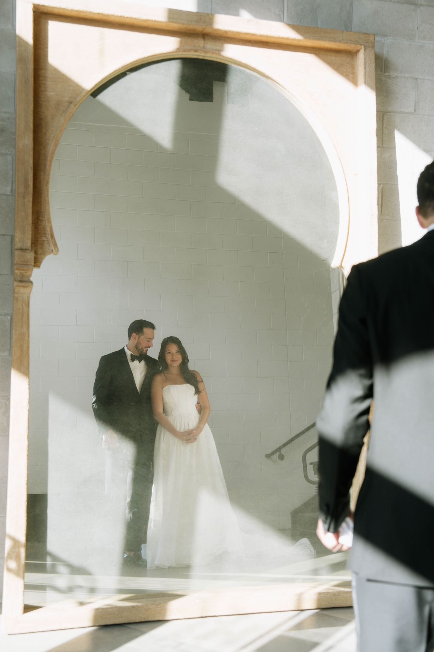 Bride and groom reflected in a large arched mirror in a sunlit elegant venue.