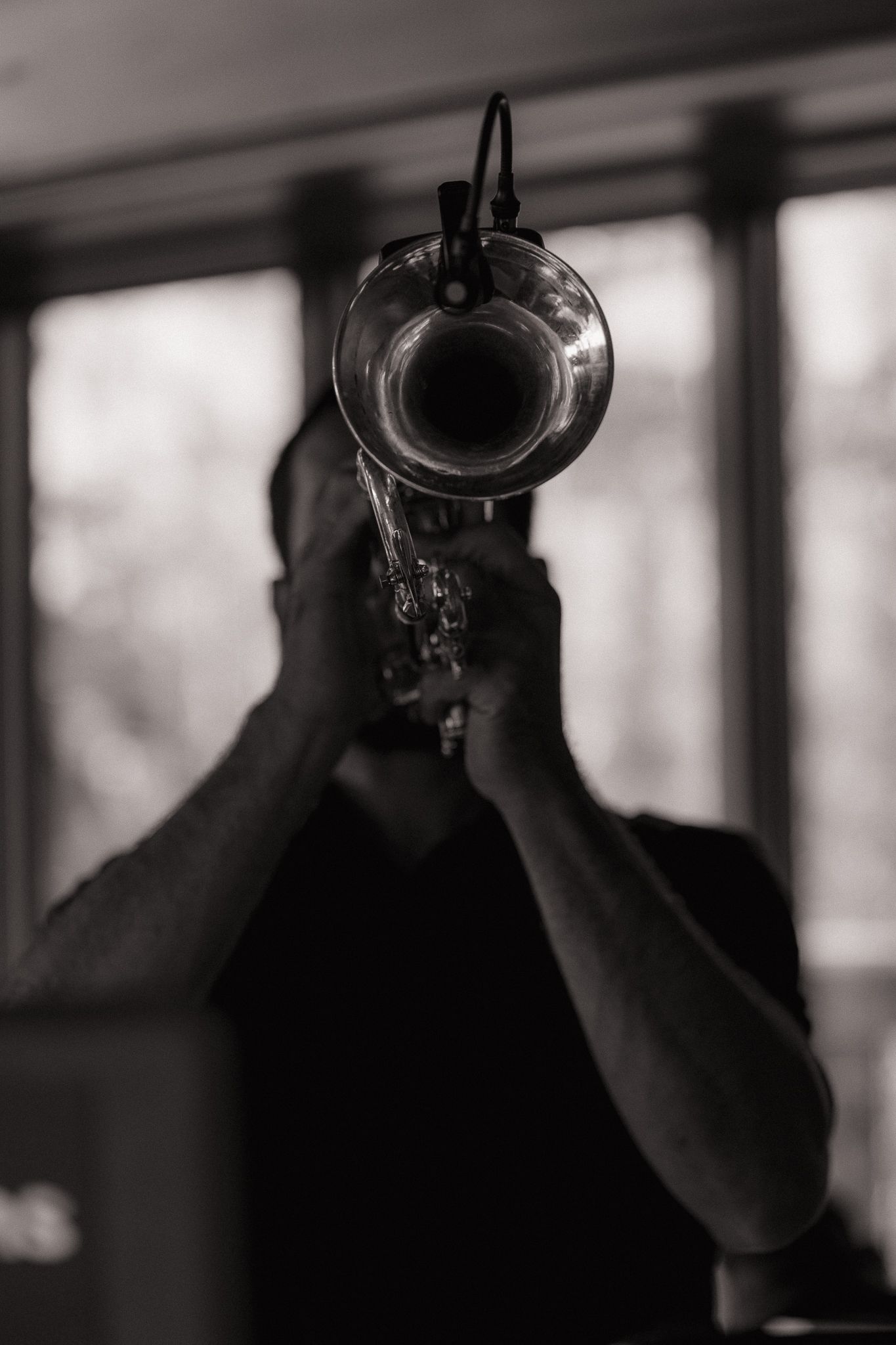 Black and white photo of a musician playing trumpet aimed at camera in moody indoor setting.