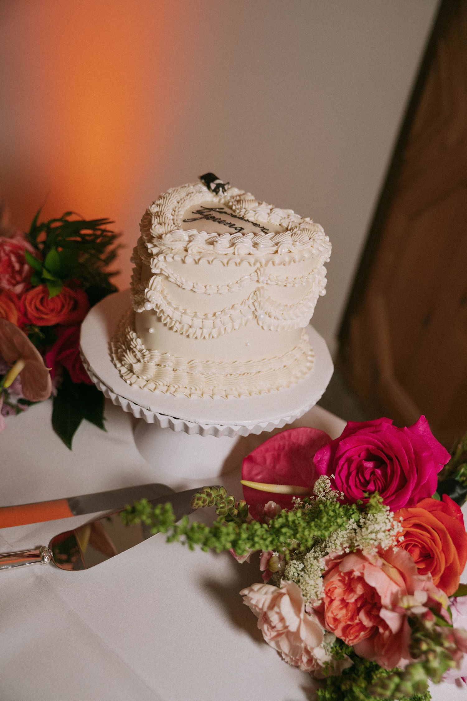 Two-tier white buttercream wedding cake with piped pearl details displayed beside vibrant pink and coral florals.