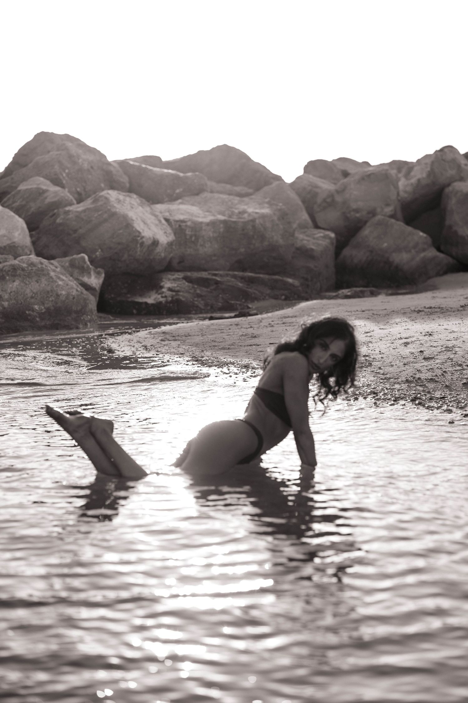 Black and white artistic photo of woman in bikini lying in shallow ocean water near rocks.