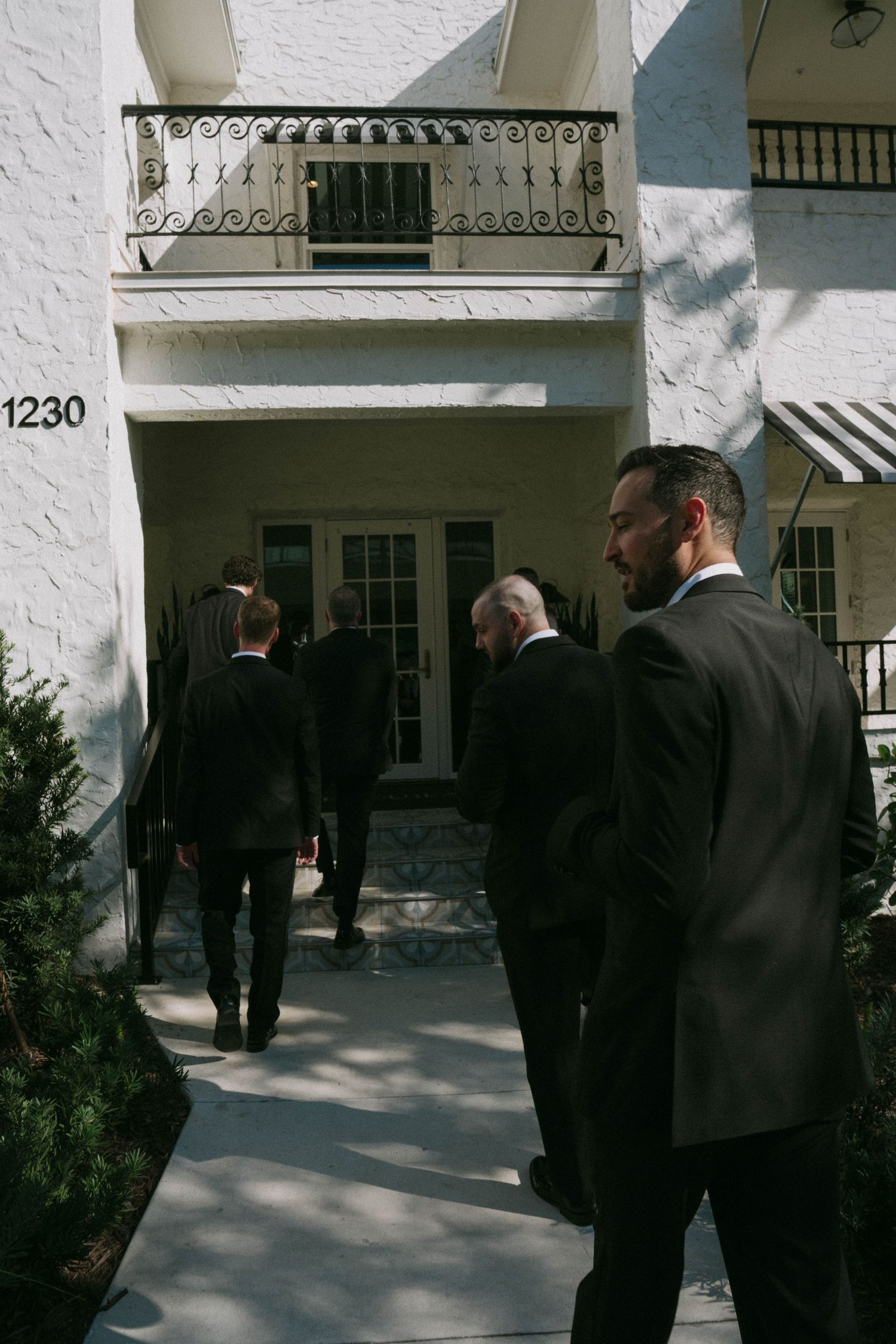 Groomsmen in dark suits walking toward a white building entrance numbered 1230.