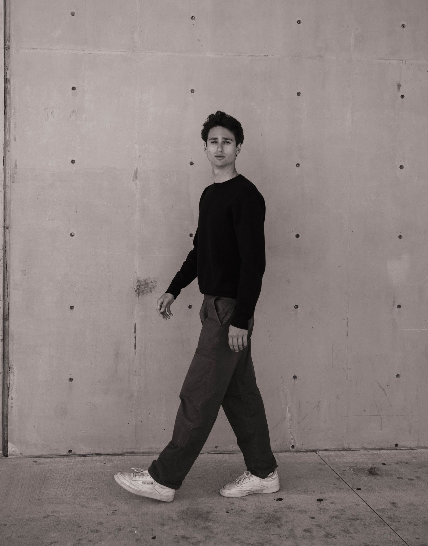 Black and white photo of a young man in a black sweater walking confidently along a concrete wall.