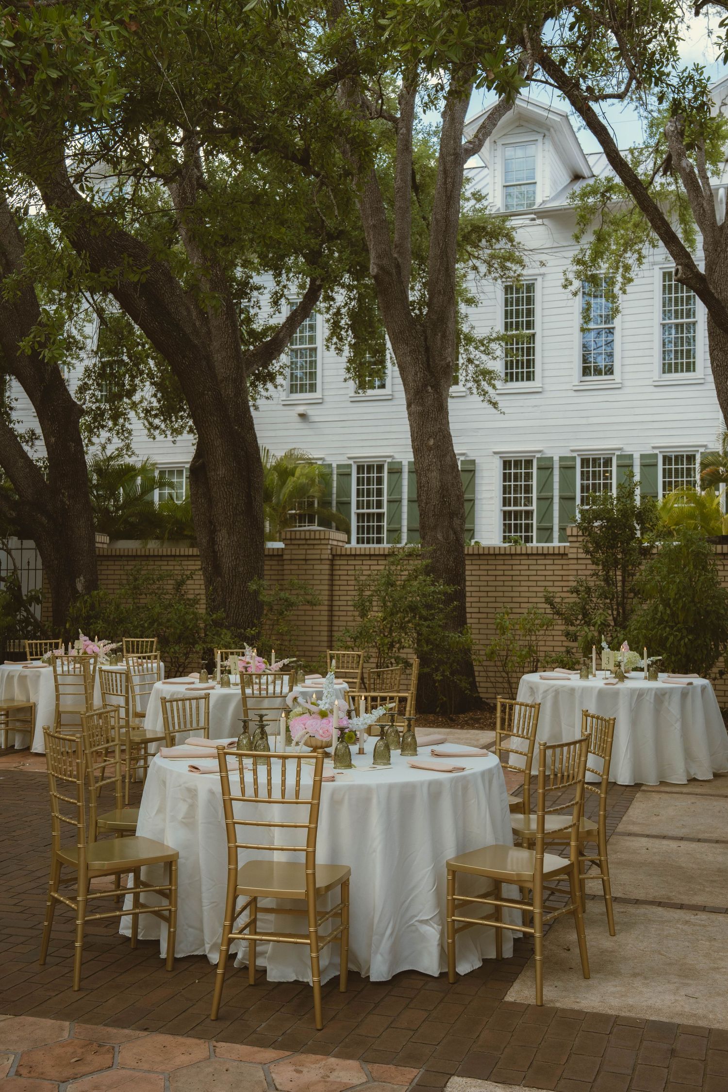 Elegant outdoor wedding reception with gold chiavari chairs and white tablecloths set beneath oak trees.