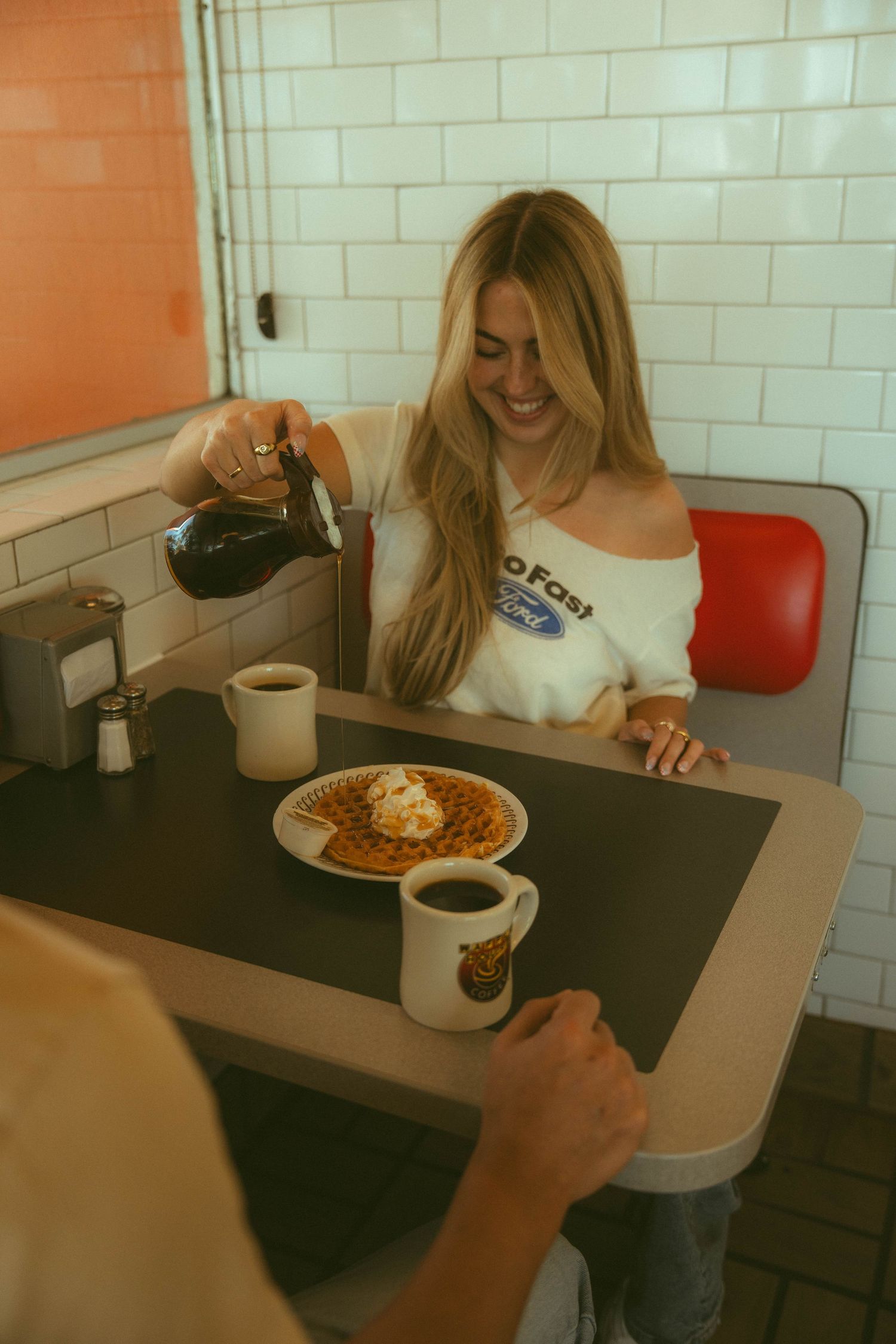 Smiling blonde woman pouring syrup on waffles at a retro diner booth with coffee.