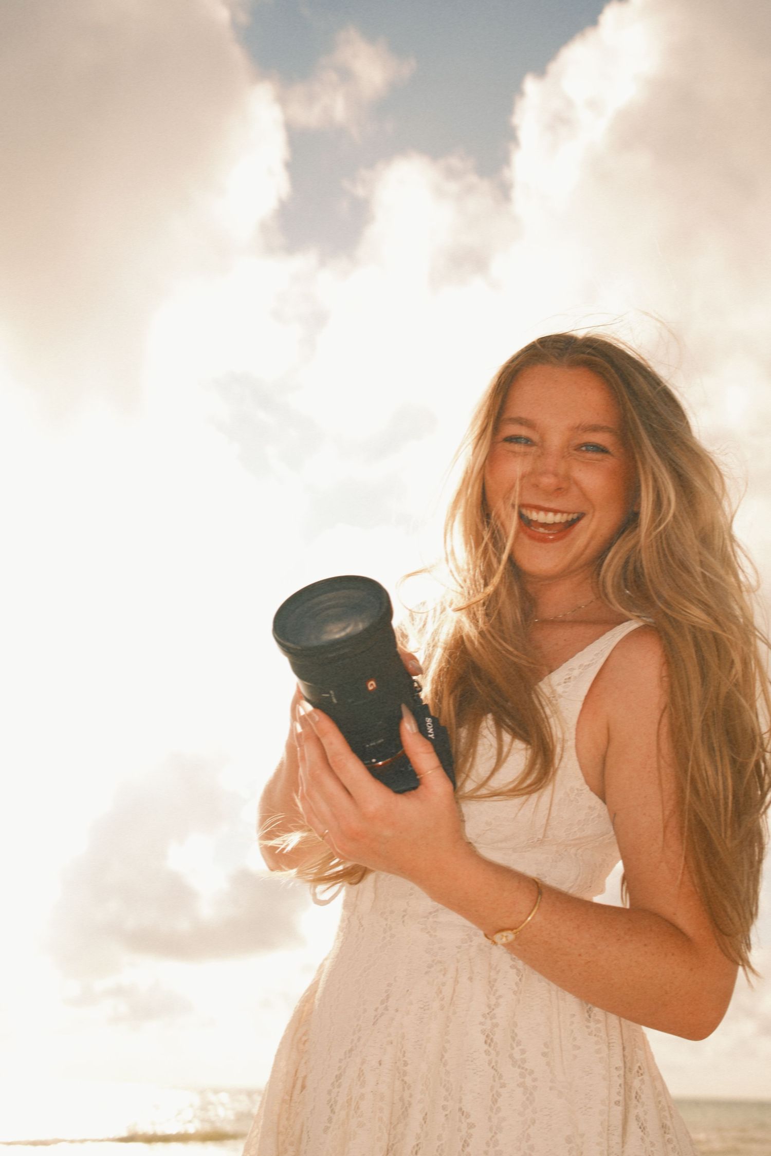 Smiling woman with long blonde hair holding a DSLR camera outdoors in a white dress with sunlit clouds behind her.