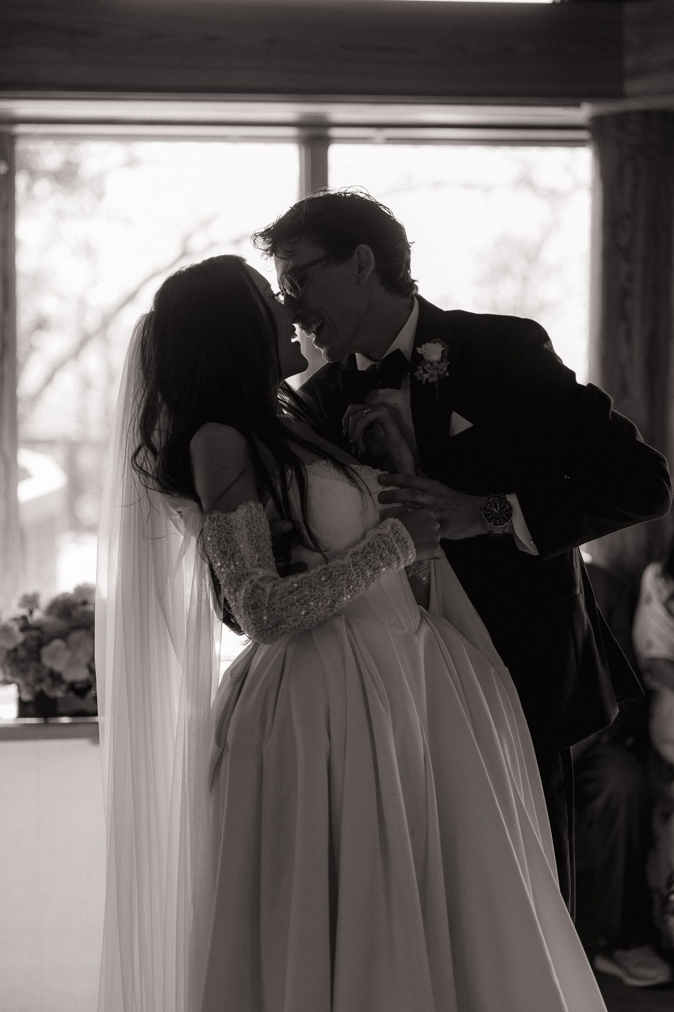 Bride and groom share a passionate kiss in black and white, bride wearing lace sleeves and flowing veil.