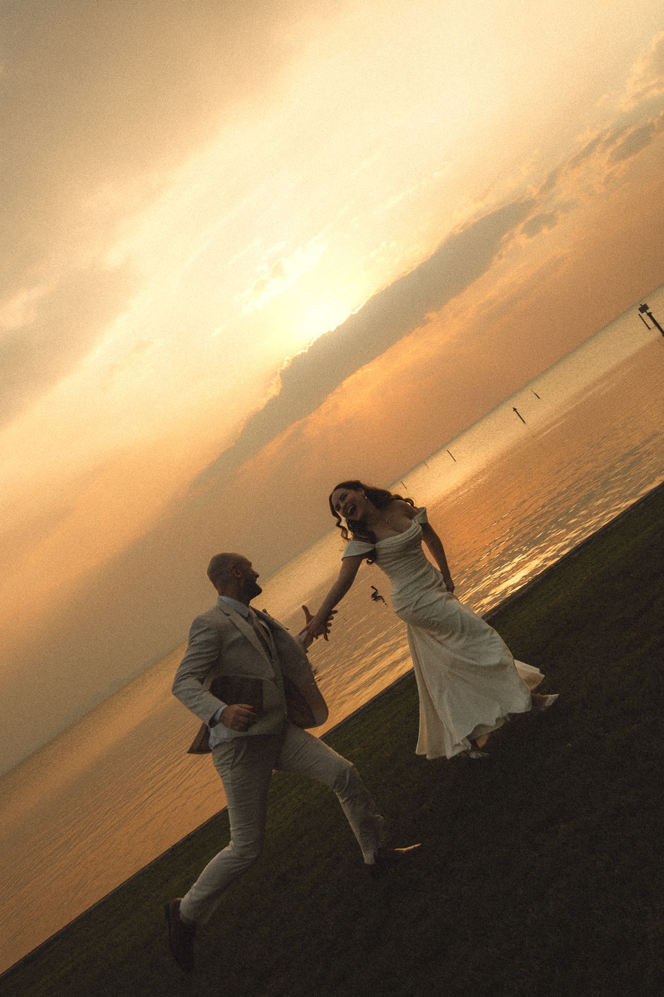 Bride and groom dancing joyfully at sunset by the waterfront, golden sky reflecting on calm waters.