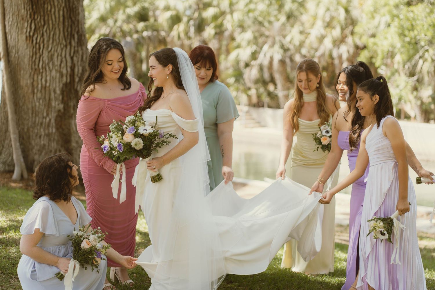 Bride surrounded by bridesmaids in colorful dresses outdoors under shaded trees on wedding day.