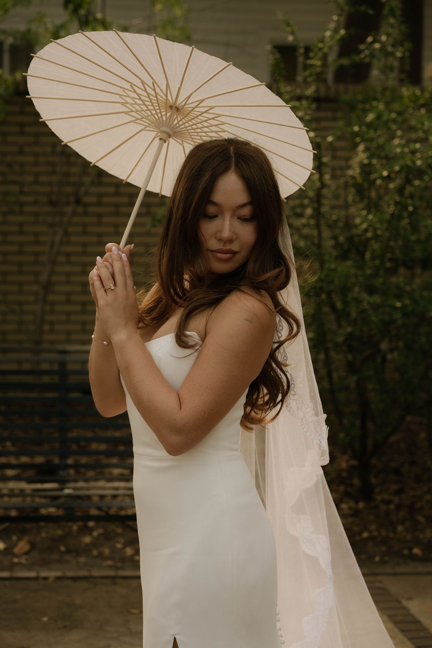 Elegant Asian bride in white gown holding parasol umbrella with lace veil in garden.