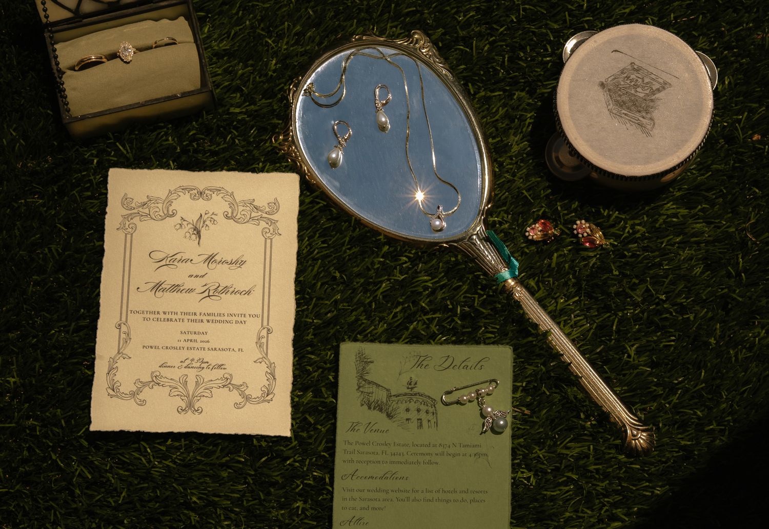 Elegant hand mirror with blue oval glass, wedding invitations, and decorative items arranged on green grass.