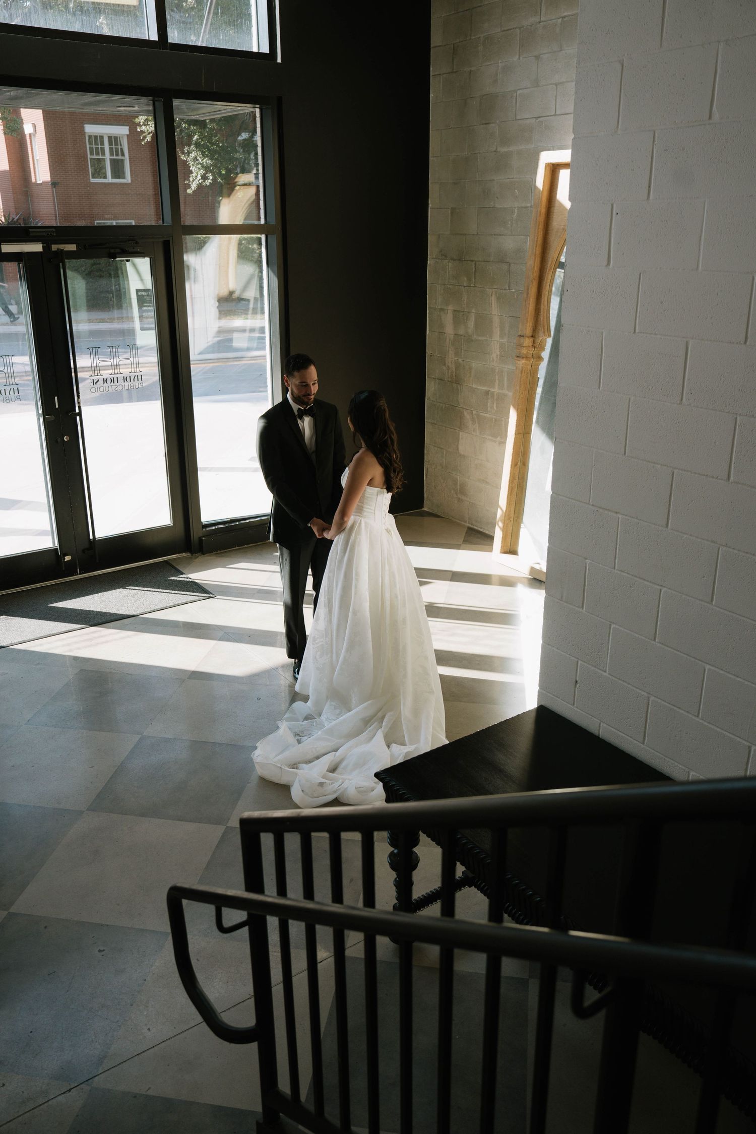 Bride in white gown and groom in black tuxedo share an intimate first look moment in a sunlit venue foyer.