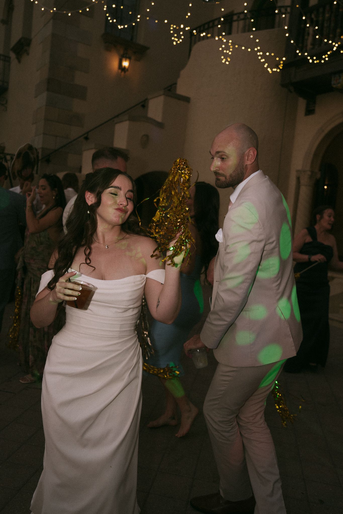 Bride in off-shoulder white gown and groom in tan suit dancing under string lights at evening reception.