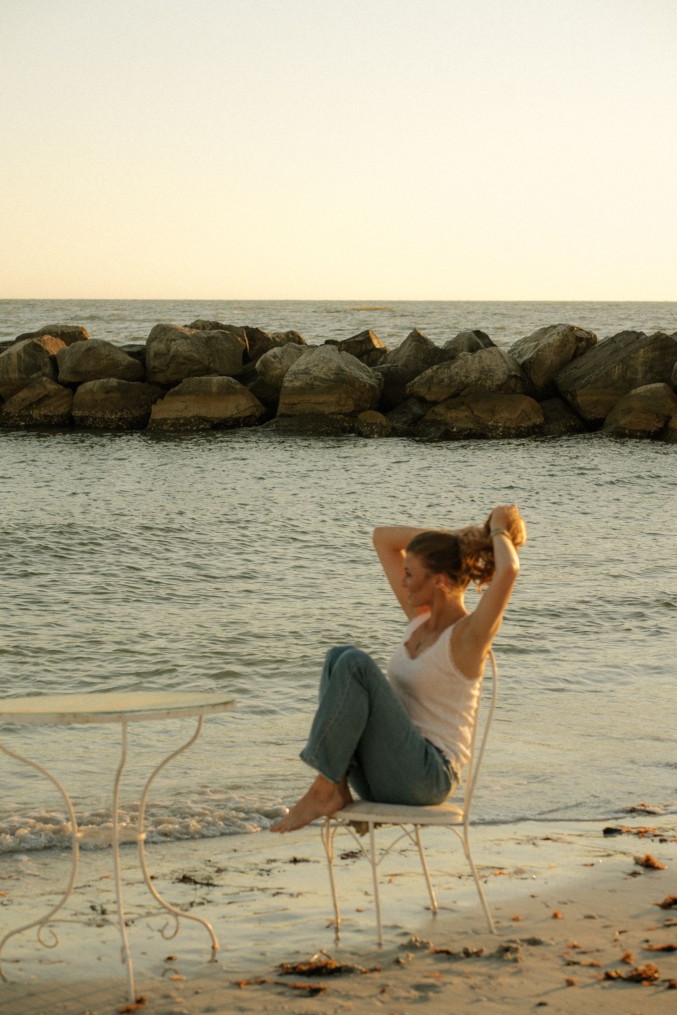 Woman relaxing on white chair at sandy beach during golden sunset with rocky breakwater and calm ocean in background.