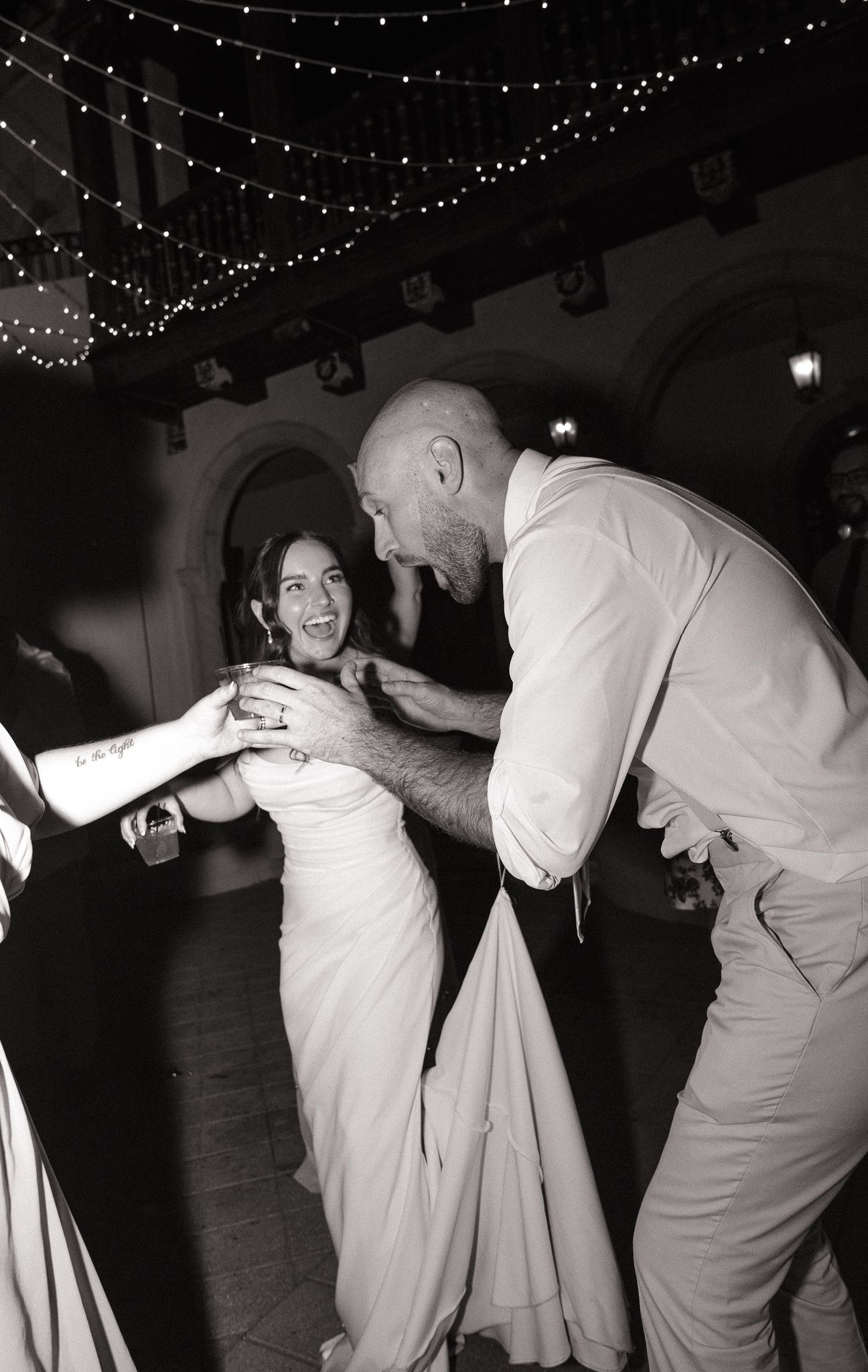 Black and white photo of a bride and groom joyfully dancing together at their wedding reception under twinkling lights.