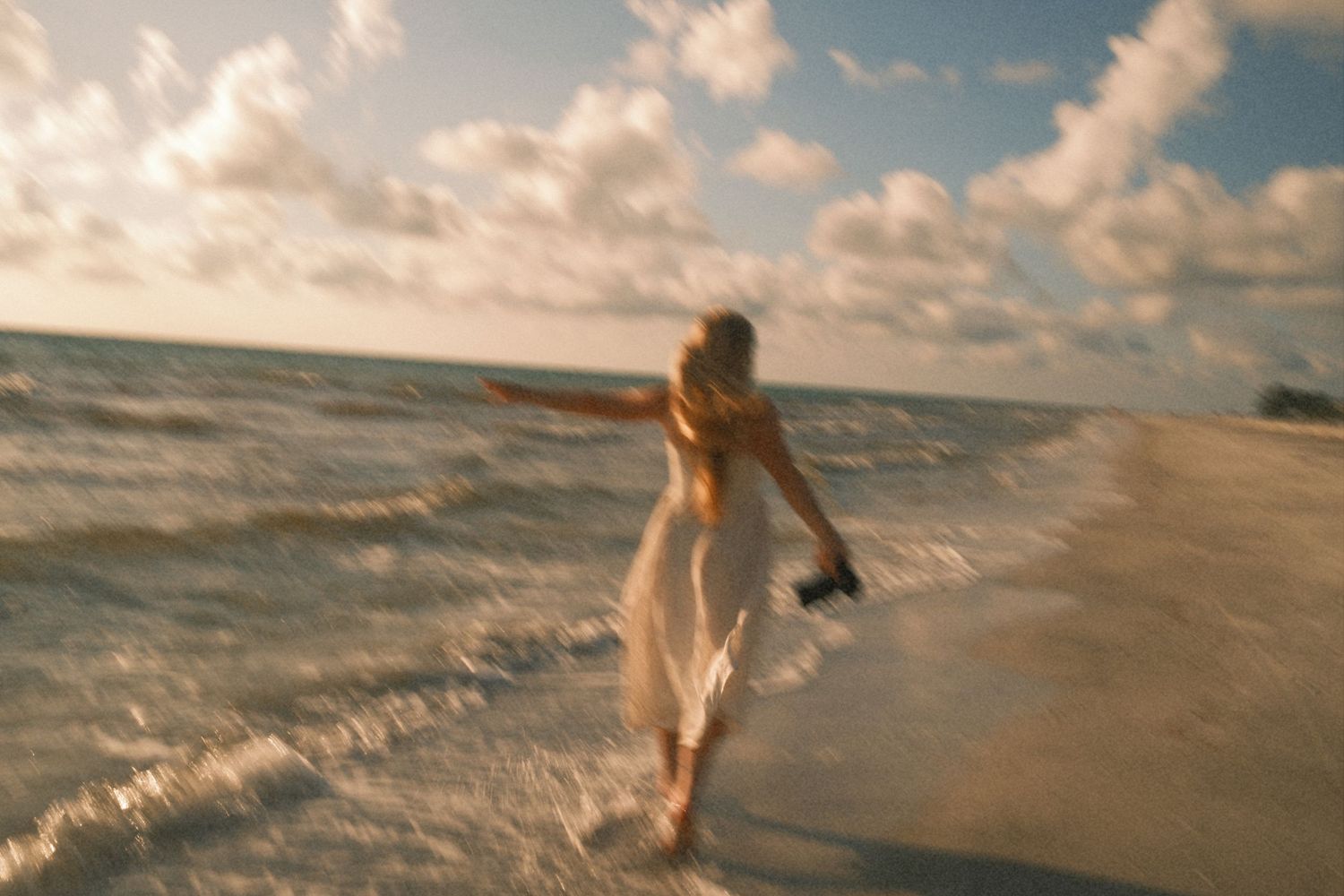A woman in a white dress runs joyfully through ocean waves on a sunny beach, holding shoes in hand.