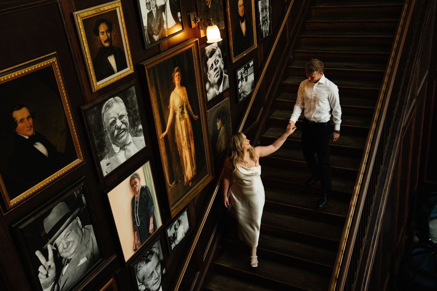 Bride and groom walk up elegant staircase lined with framed portraits in a dimly lit venue.