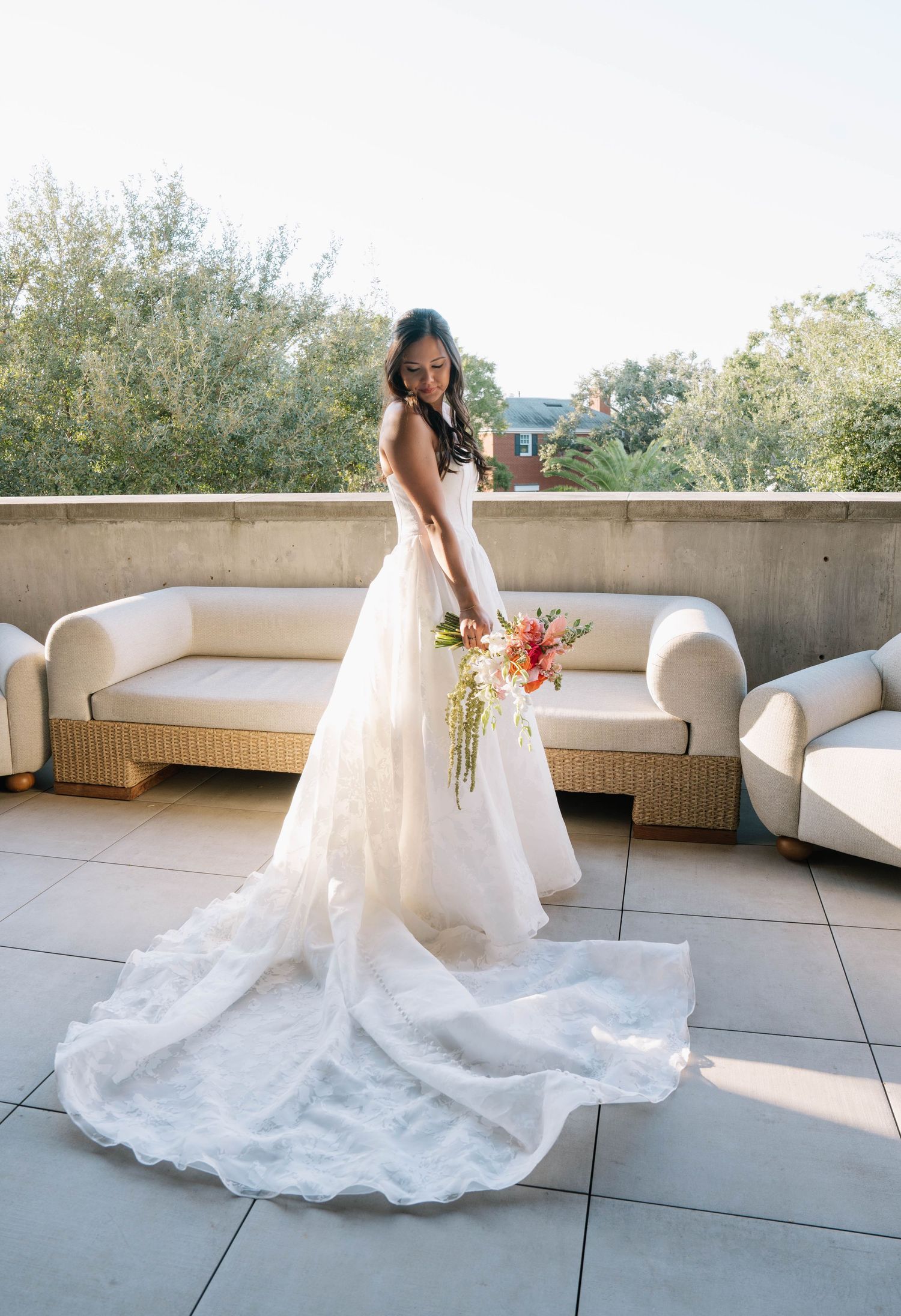 Elegant bride in a lace ball gown with a long train holds a colorful bouquet on a sunny rooftop terrace.