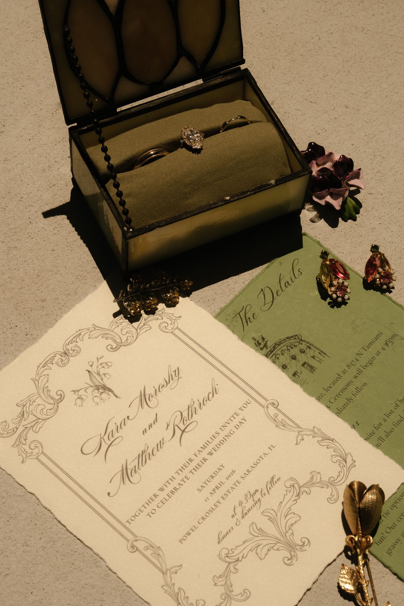 Elegant wedding invitation suite with rings in a glass box, floral details, and a green details card on textured surface.