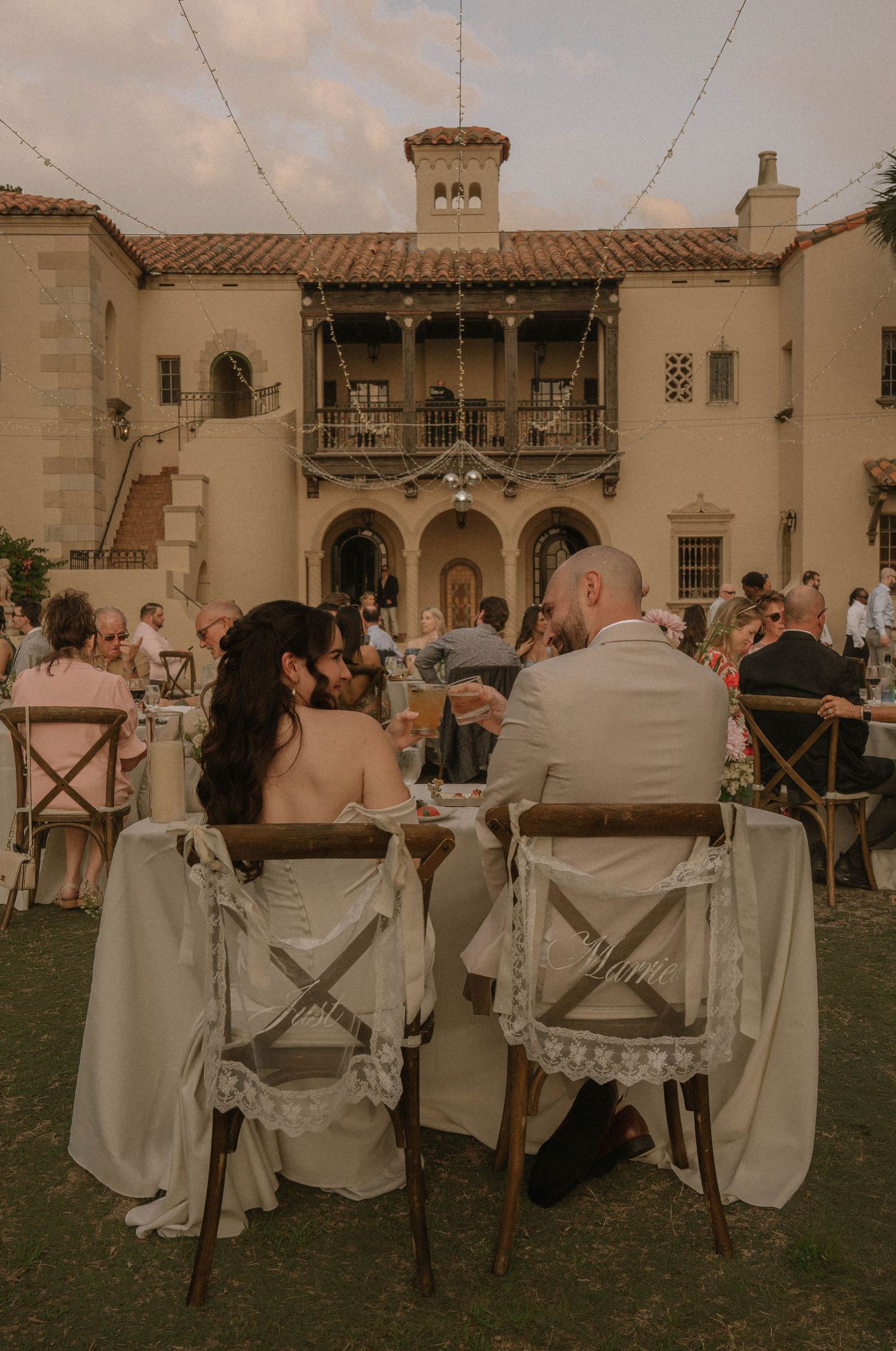 Bride and groom sit at sweetheart table during outdoor wedding reception at elegant Spanish-style mansion.