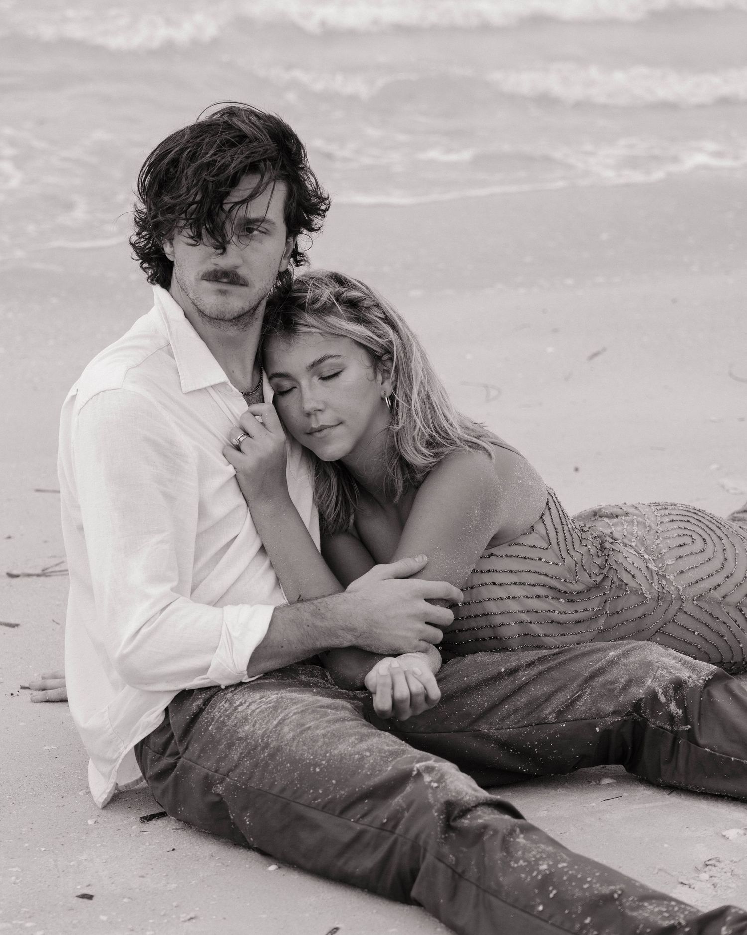 Black and white photo of a couple sitting on a beach, embracing tenderly with ocean waves behind them.