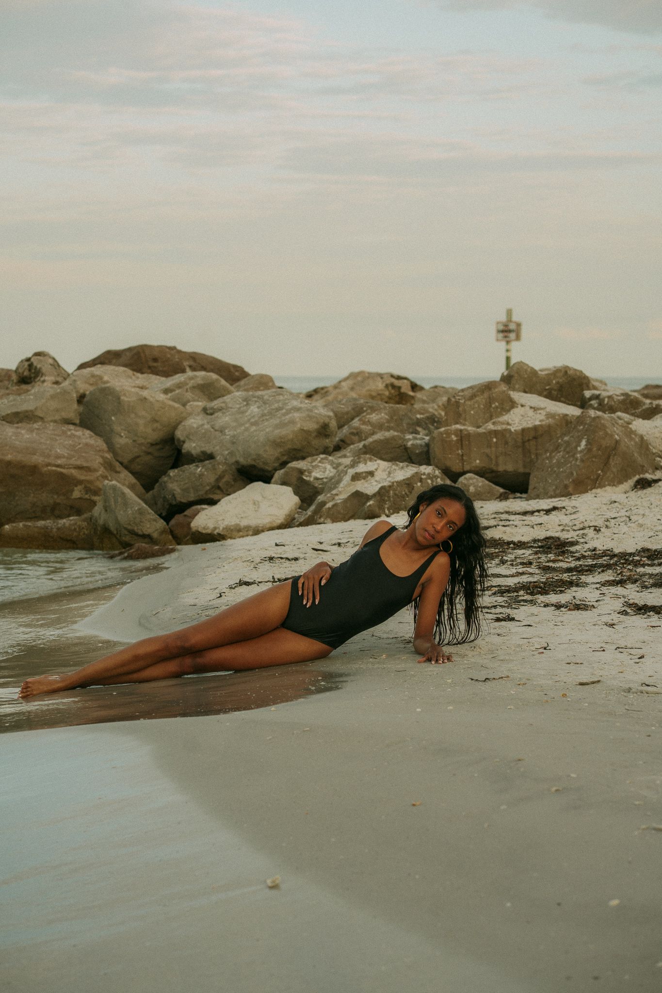 Woman in black swimsuit reclines on sandy beach near rocky shore in a relaxed, elegant pose at dusk.