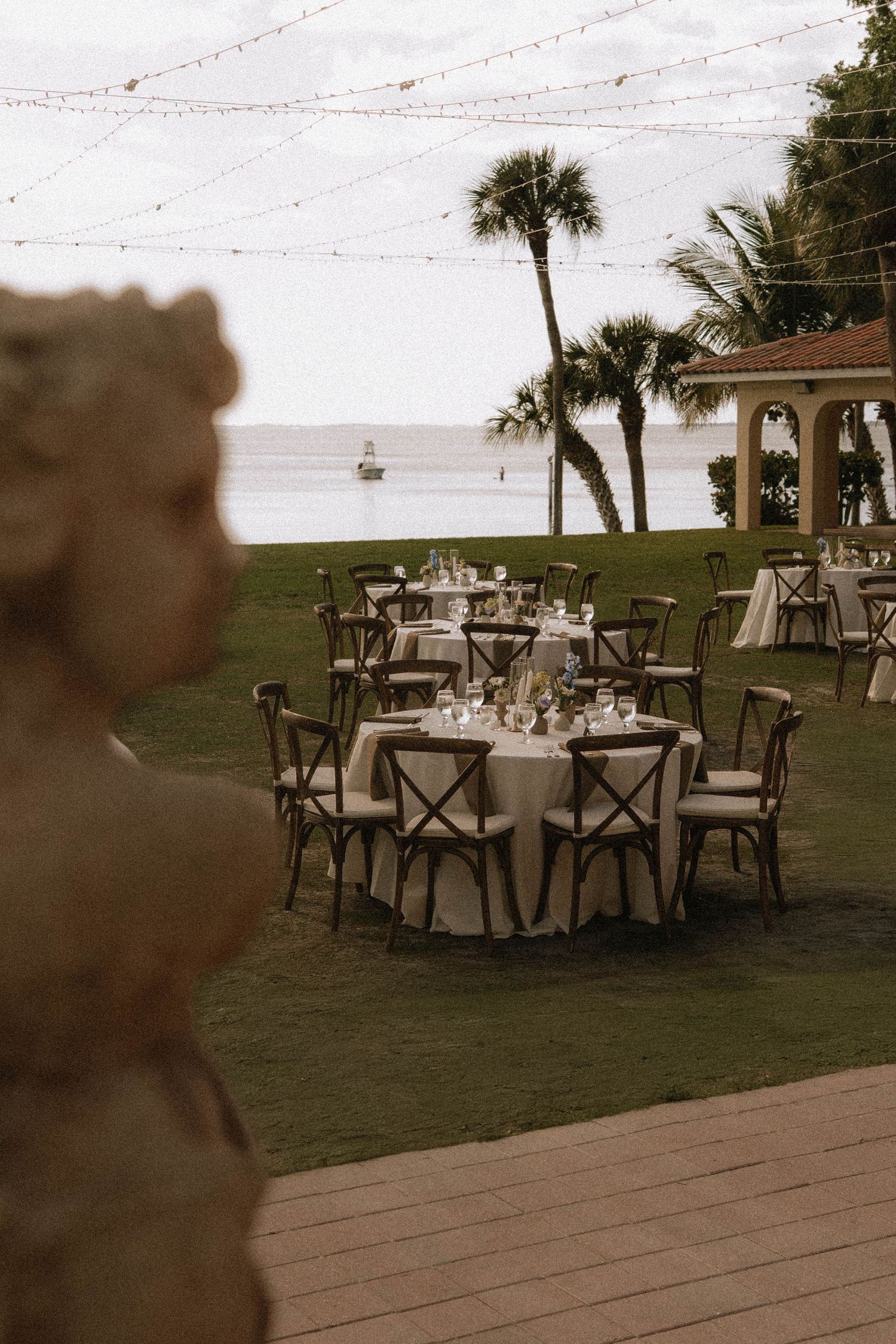 Elegant outdoor wedding reception setup with round tables, string lights, palm trees, and ocean view.