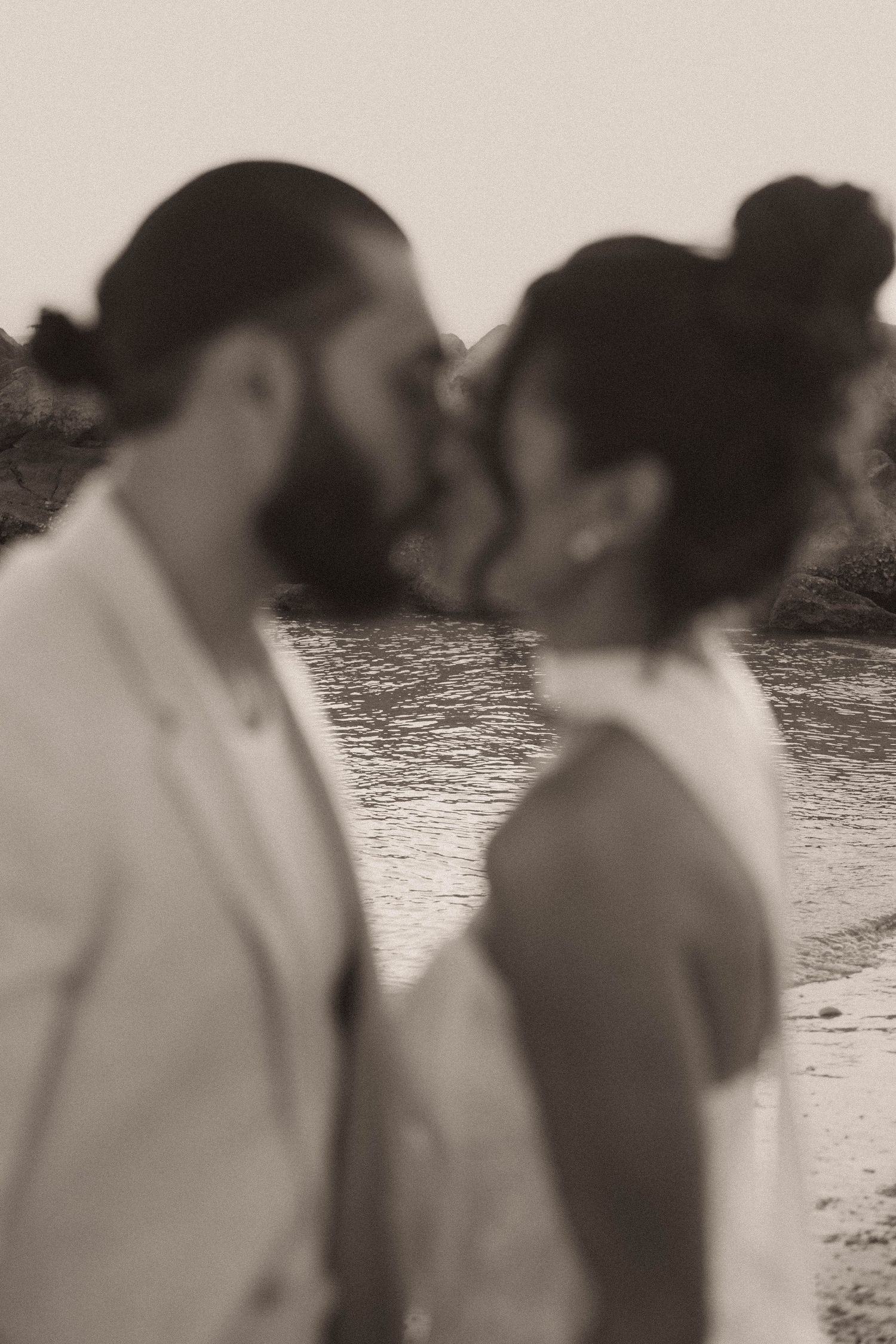 Soft-focus sepia image of bearded groom and bride sharing a kiss on a sunlit beach.