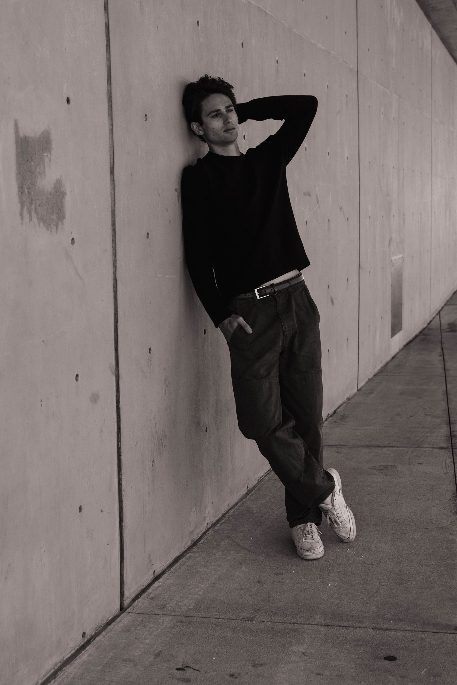 Stylish young man in black sweater leaning against concrete wall in black and white urban portrait.