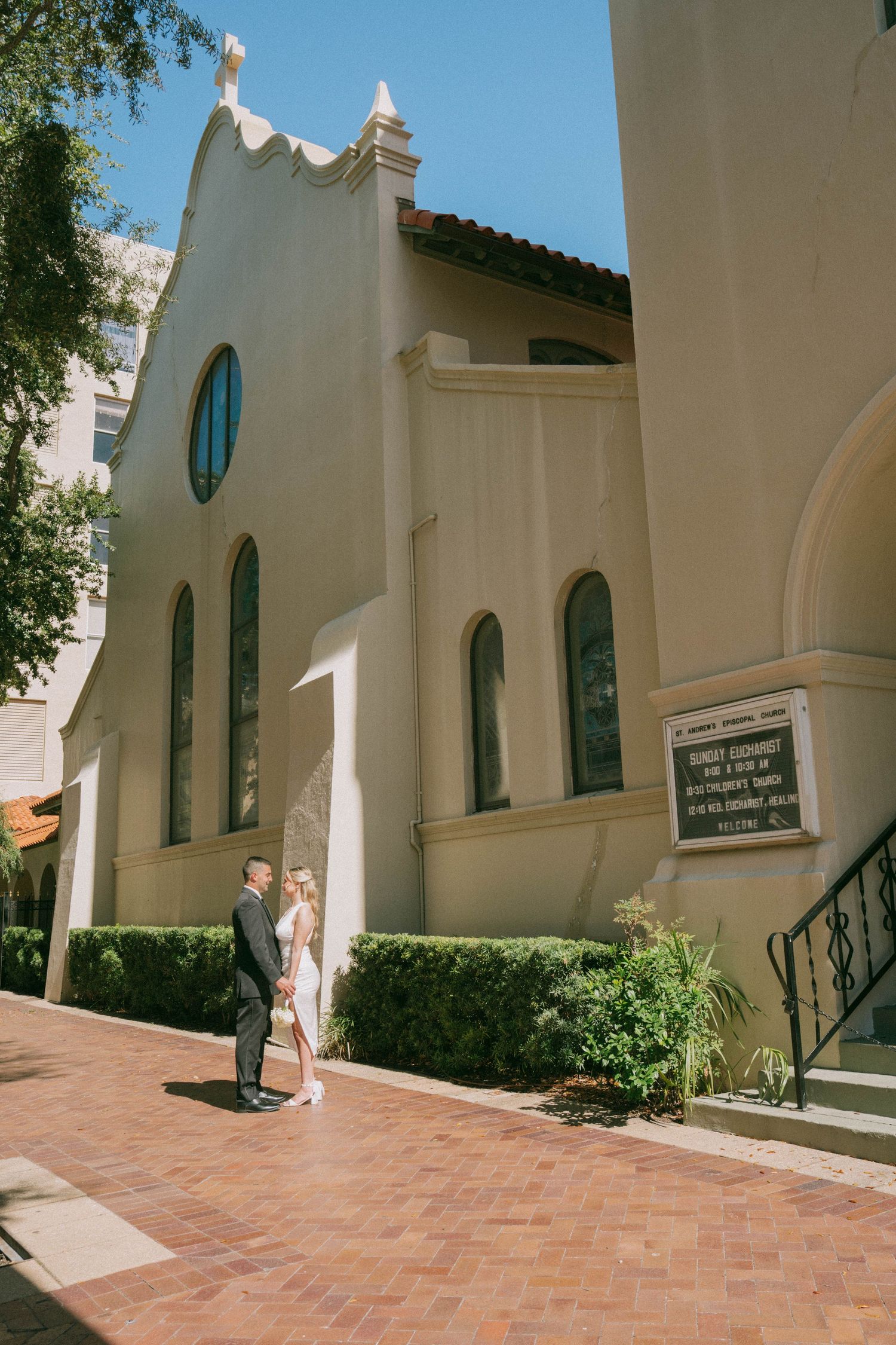 Bride and groom embracing on brick pathway outside Spanish-style church in bright sunlight.
