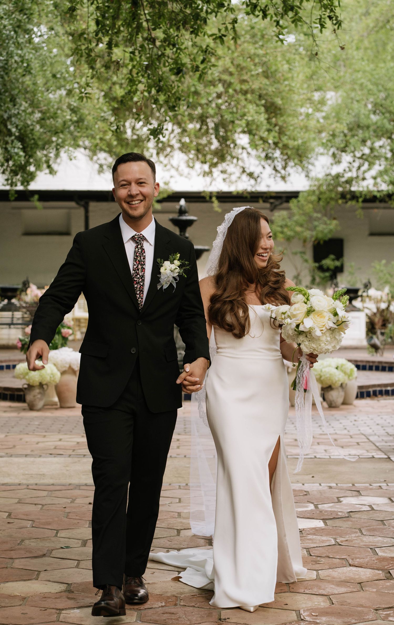 Smiling newlyweds walk hand-in-hand in a courtyard, groom in black suit, bride in white fitted gown with bouquet.