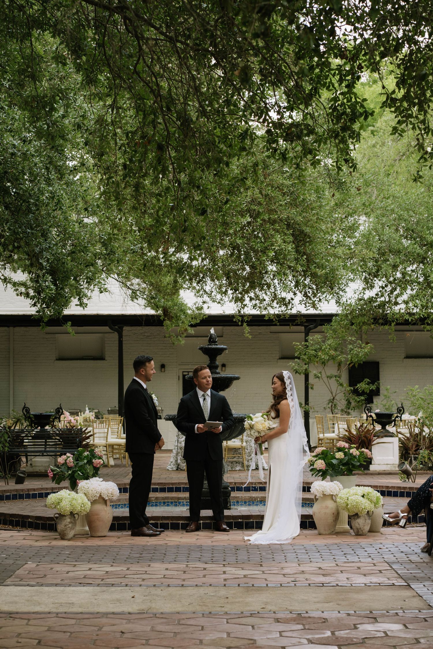 Bride and groom exchange vows outdoors near fountain under lush green trees.