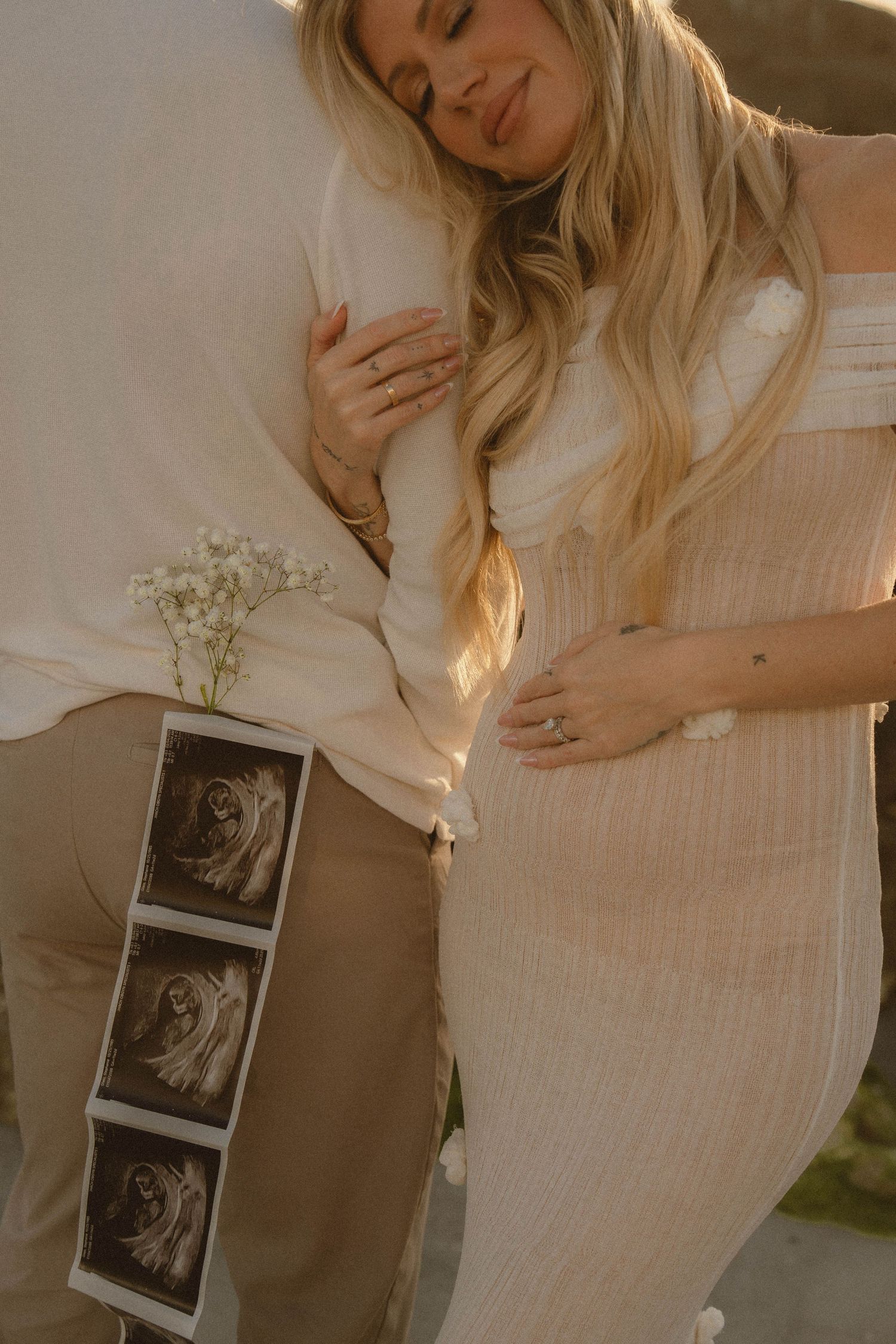 Pregnant woman in beige dress embracing partner outdoors holding ultrasound photos and baby breath flowers.